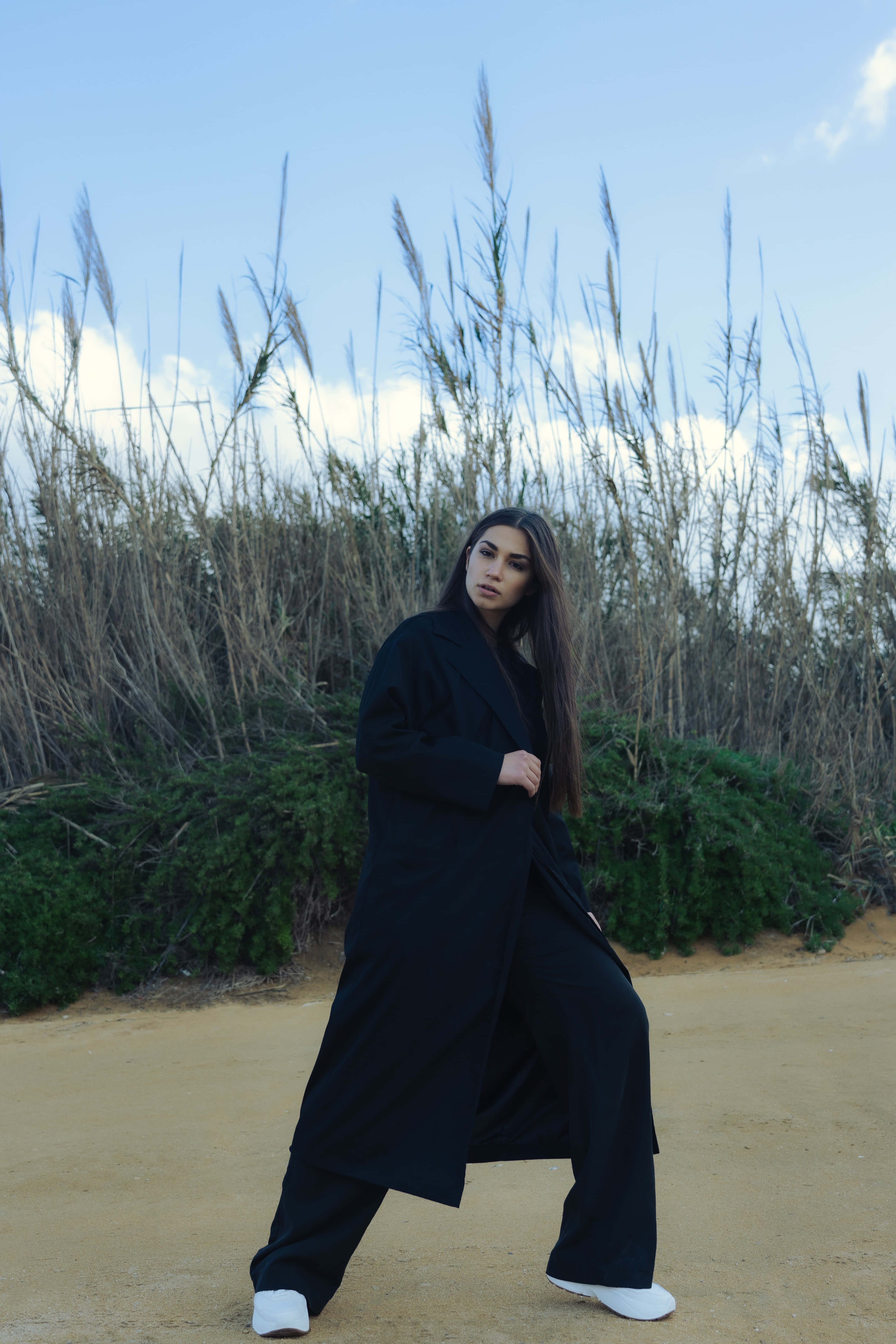 Navy Blue Coat 桌布 壁紙 — 一位身穿黑色外套和白色鞋子的女性自信地站在一條泥濘小徑上，周圍是高高的蘆葦，頭頂是清澈的藍天。