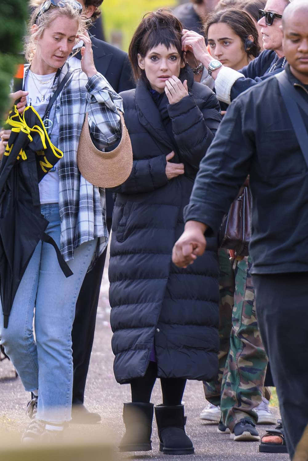  壁紙 — 一名身穿黑色羽絨外套和靴子的女性，在晴朗的日子裡穿過戶外的人群。