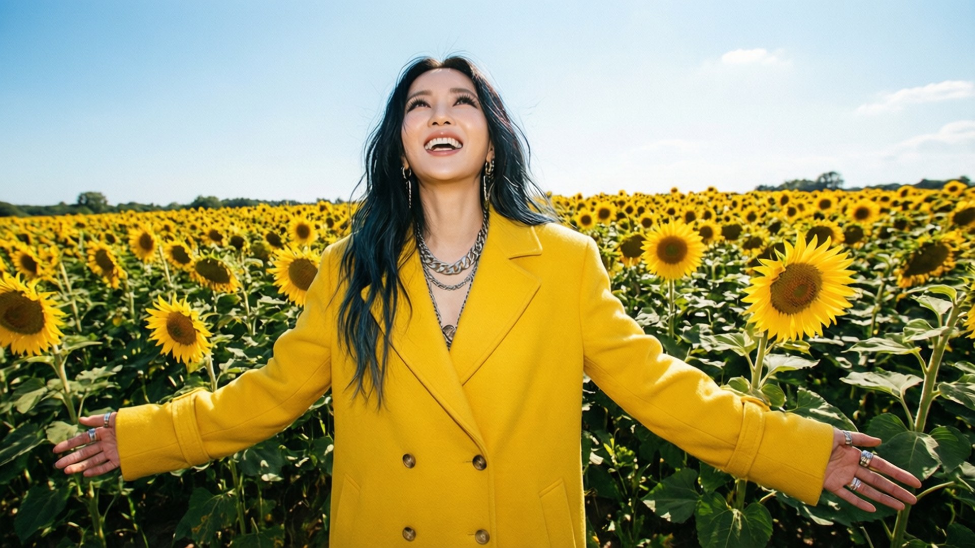 Sunflower Fields 壁紙 — 一位身穿亮黃色外套的女性在晴朗的藍天下，在一片廣闊的向日葵田中張開雙臂，喜悅地站立著。