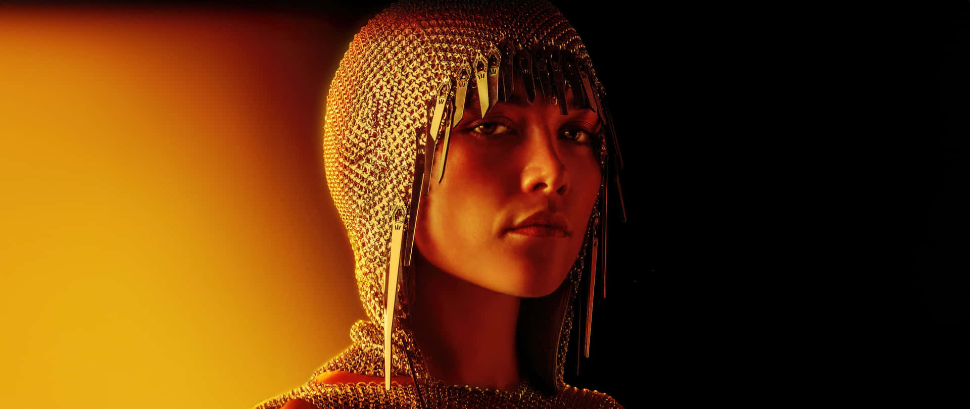 壁紙 — 一位戴著金色鎖子甲頭罩的女性在溫暖的橙色燈光下，於深色背景中向前凝視。