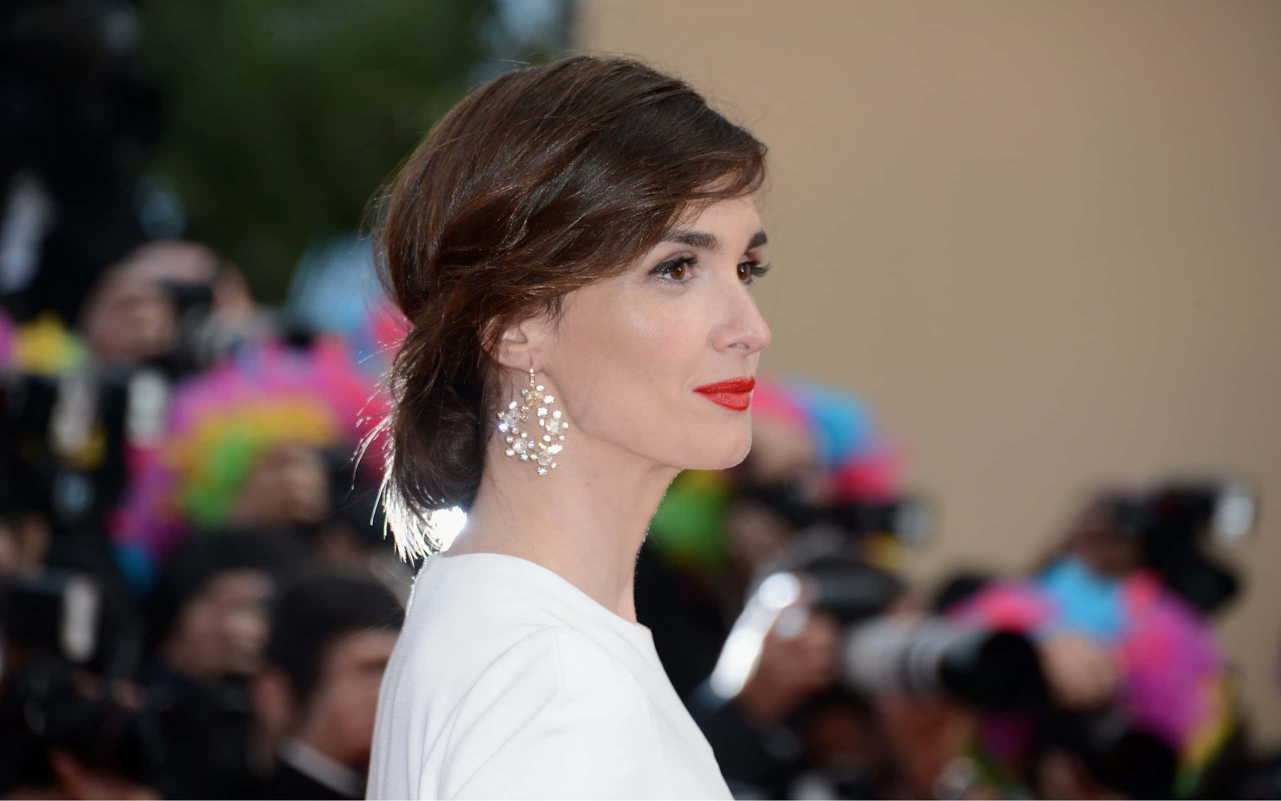 Paz Vega 桌布 壁紙 — 一位側面女士身穿白色上衣，戴著閃亮耳環，背景是模糊的攝影師人群。