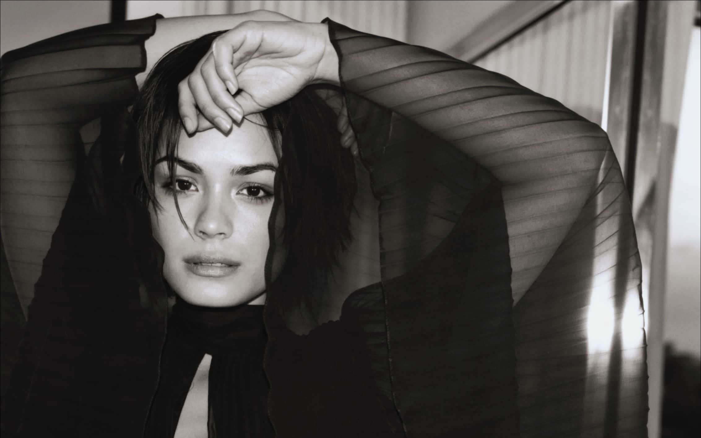 Shannyn Sossamon 桌布 壁紙 — 一位深色頭髮的女性穿著透視黑色上衣，一手放在額頭上，直視鏡頭，營造出憂鬱的黑白肖像。