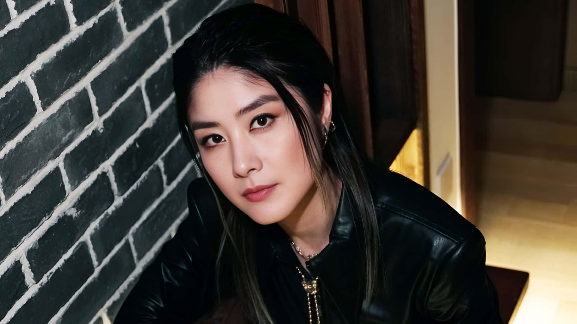  壁紙 — 一位深色頭髮、眼神銳利的女士，在溫暖的室內燈光下，穿著黑色皮夾克靠著黑色磚牆。