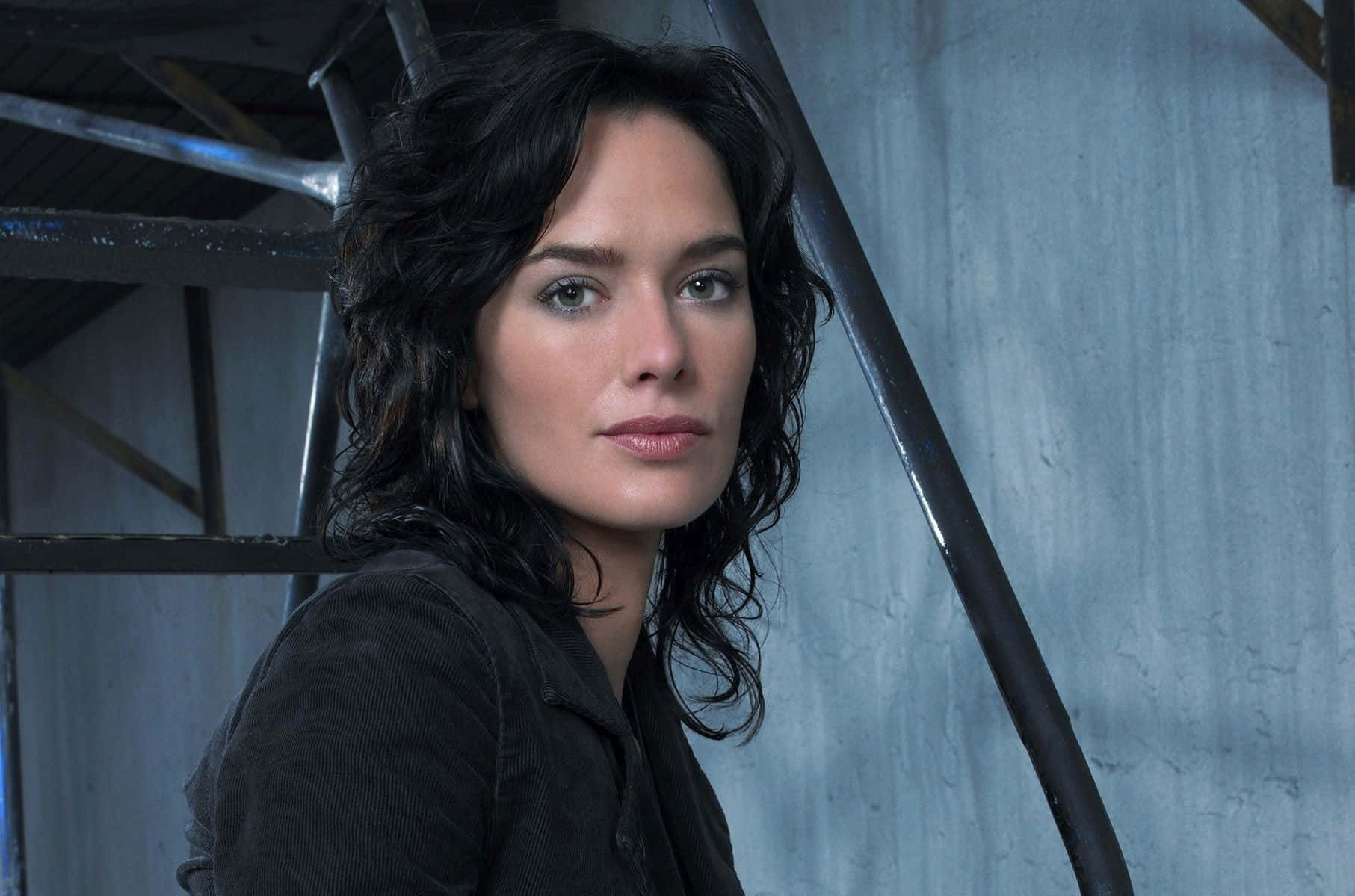 Lena Headey 桌布 壁紙 — 一位擁有深色波浪髮的女性在紋理藍色工業牆和金屬樓梯背景下直視鏡頭。