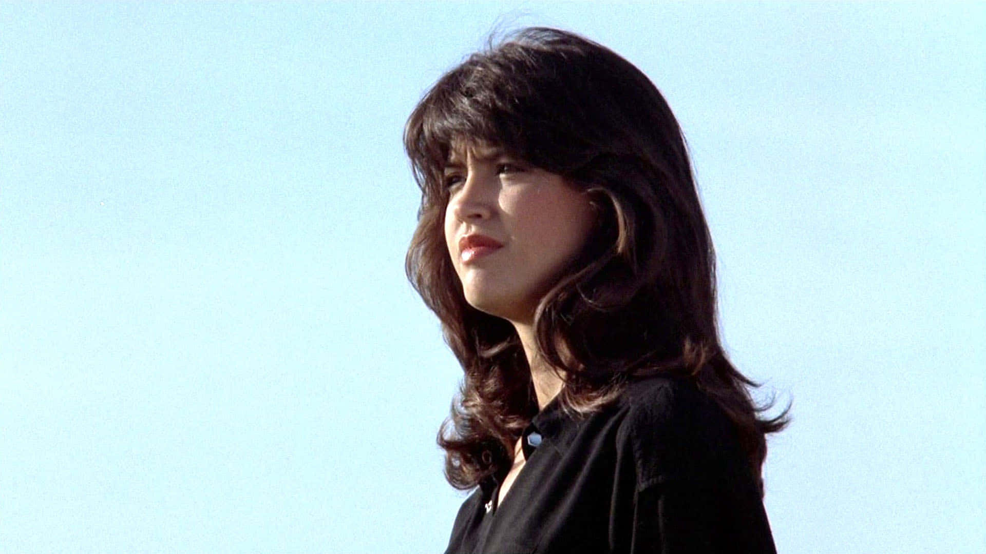 Phoebe Cates 桌布 壁紙 — 一位擁有深色波浪長髮的女性，在柔和日光下，若有所思地凝視著清澈的藍天。