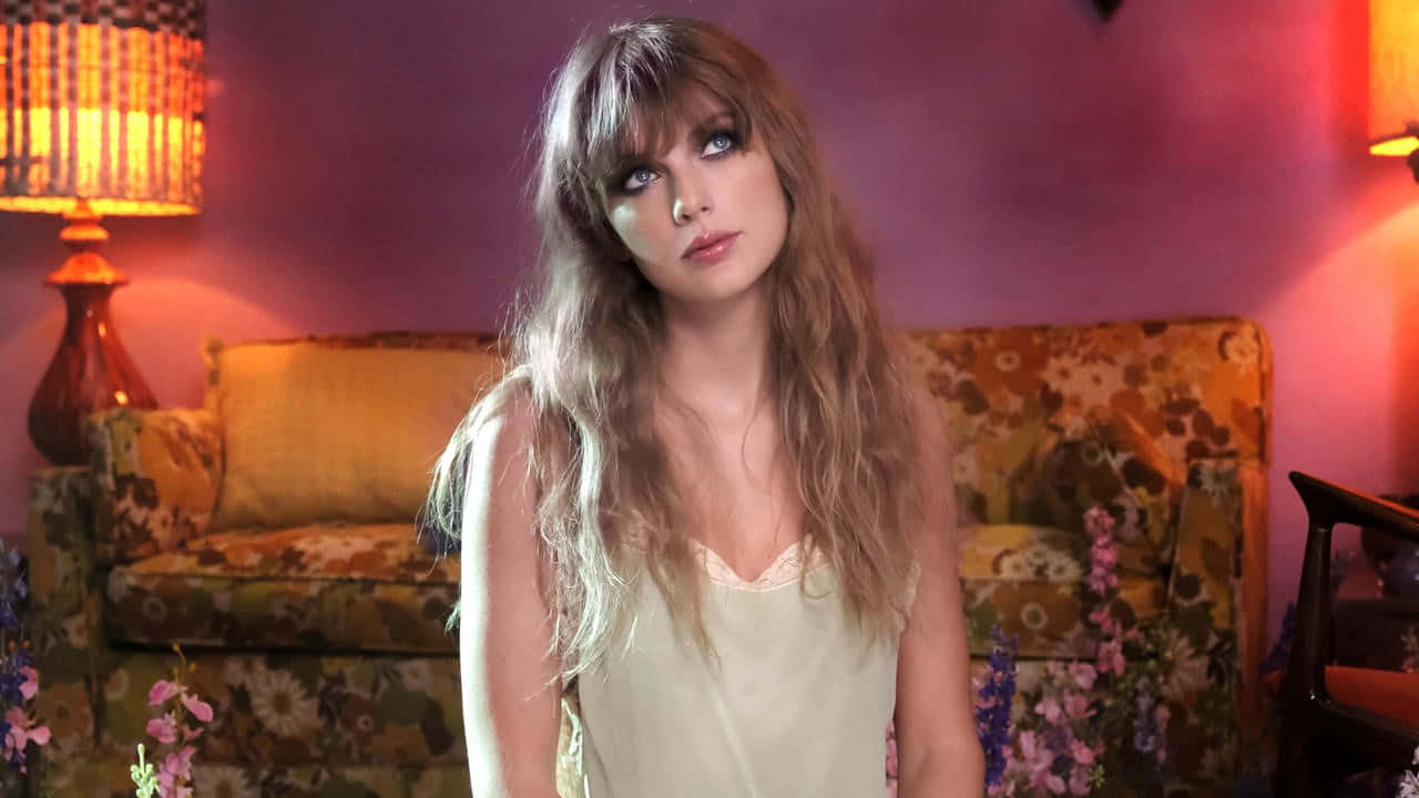Taylor Swift Desktop 桌布 壁紙 — 一位長金髮的女性坐在一個光線溫暖的房間裡，房間裡有花卉家具和發出柔和陰影的發光燈具。
