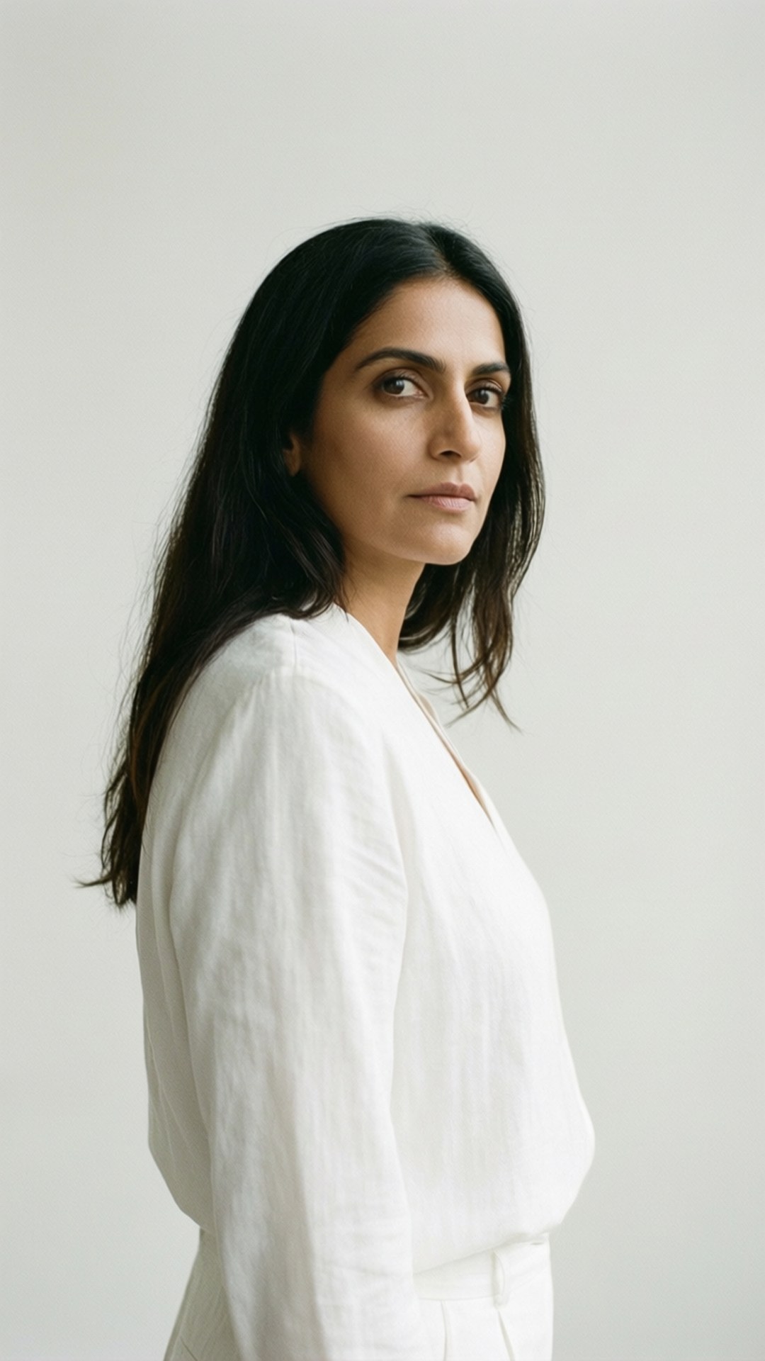 Minimalist Elegance 壁紙 — 一位長黑髮的女性穿著白色亞麻襯衫，在純淺灰色背景前直視鏡頭。
