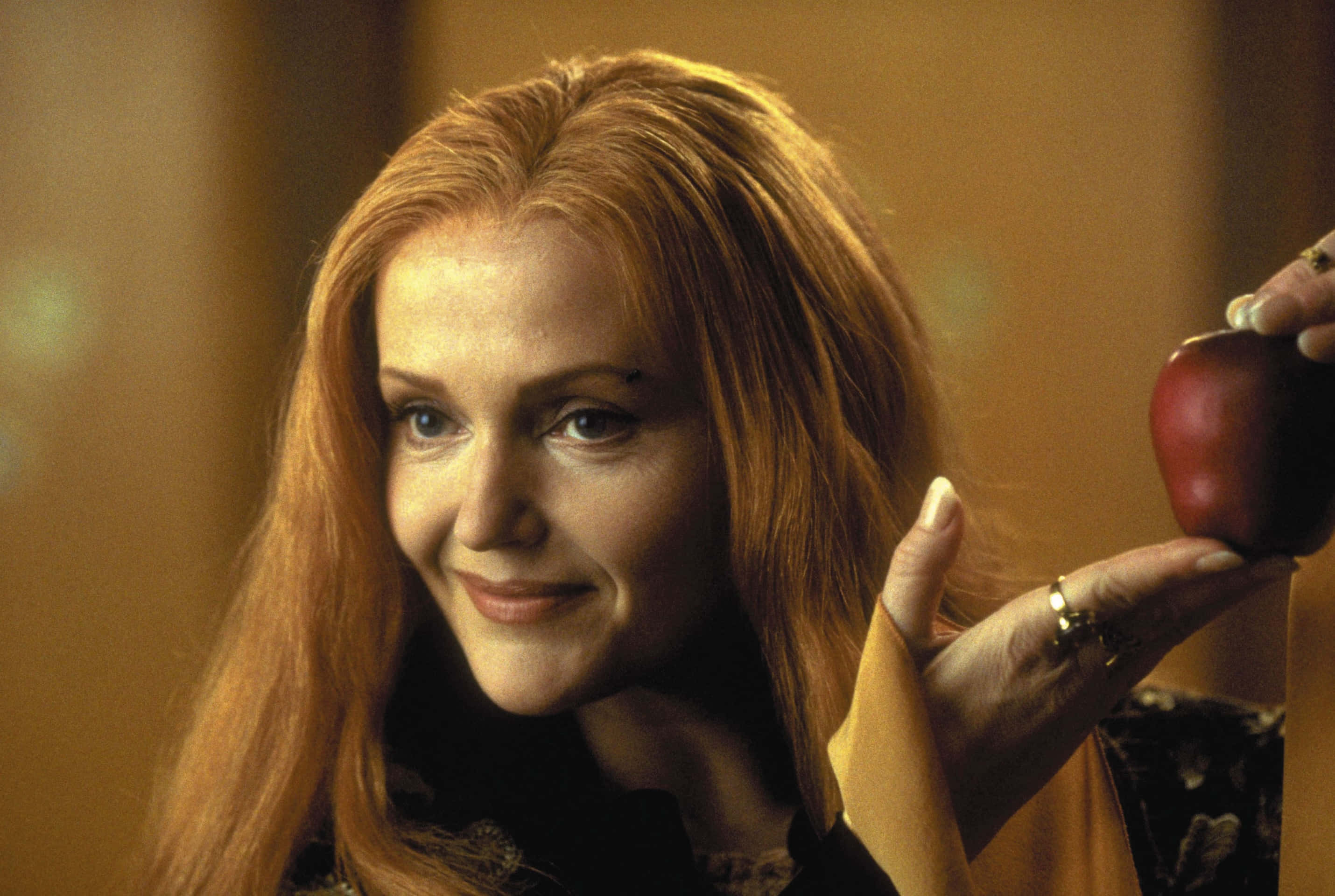 Miranda Richardson 桌布 壁紙 — 一位長紅髮的女性在溫暖燈光的室內環境中微笑著拿著一個紅蘋果。