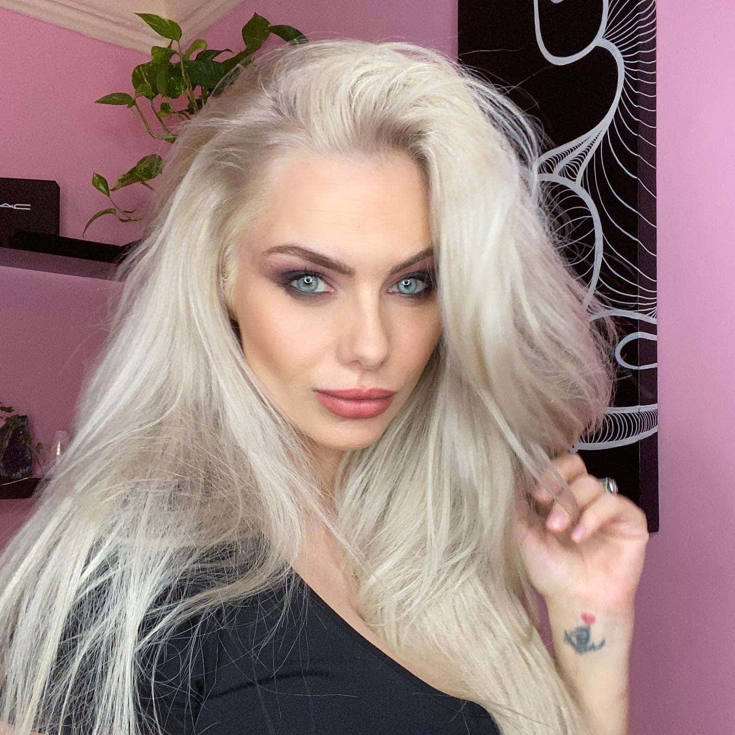 Jessica Jane Clement 桌布 壁紙 — 一位擁有鉑金色頭髮和引人注目的藍眼睛的女性，在一個有綠色植物和黑色牆壁藝術的粉紅色房間裡擺姿勢。