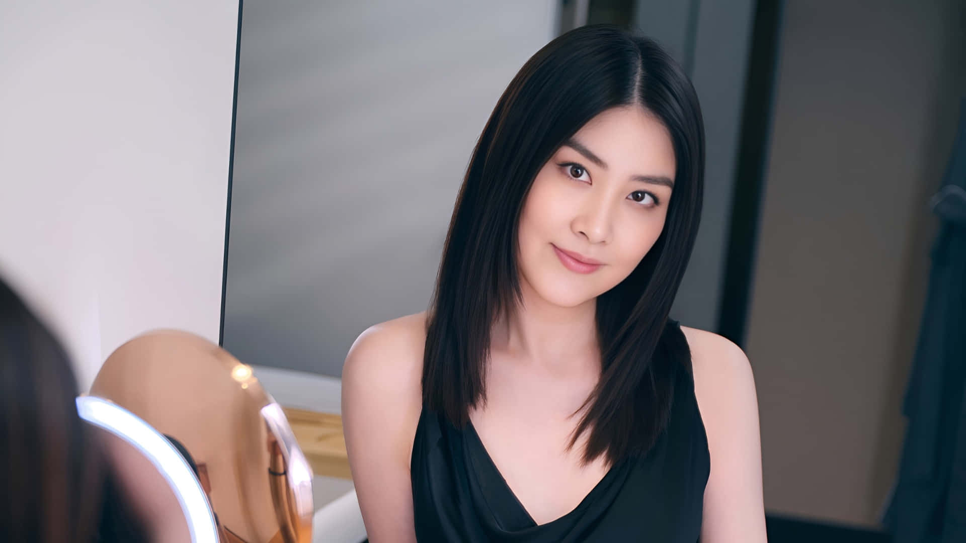  壁紙 — 一位擁有黑色直髮的女性，在帶有環形燈的鏡子前看著自己的倒影，溫柔地微笑。