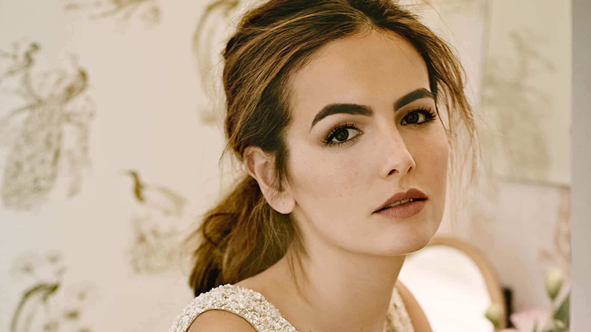 Camilla Belle 桌布 壁紙 — 一位擁有時尚髮型和優雅妝容的女性，在精緻圖案的壁紙背景下溫柔凝視。