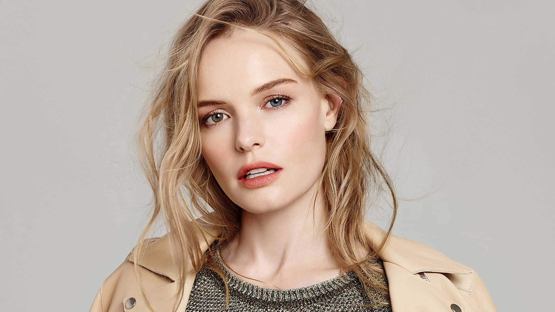 Kate Bosworth 桌布 壁紙 — 一位擁有凌亂金髮和引人注目的藍眼睛的女性，在灰色中性背景前，穿著米色風衣搭配質感毛衣，直視鏡頭。