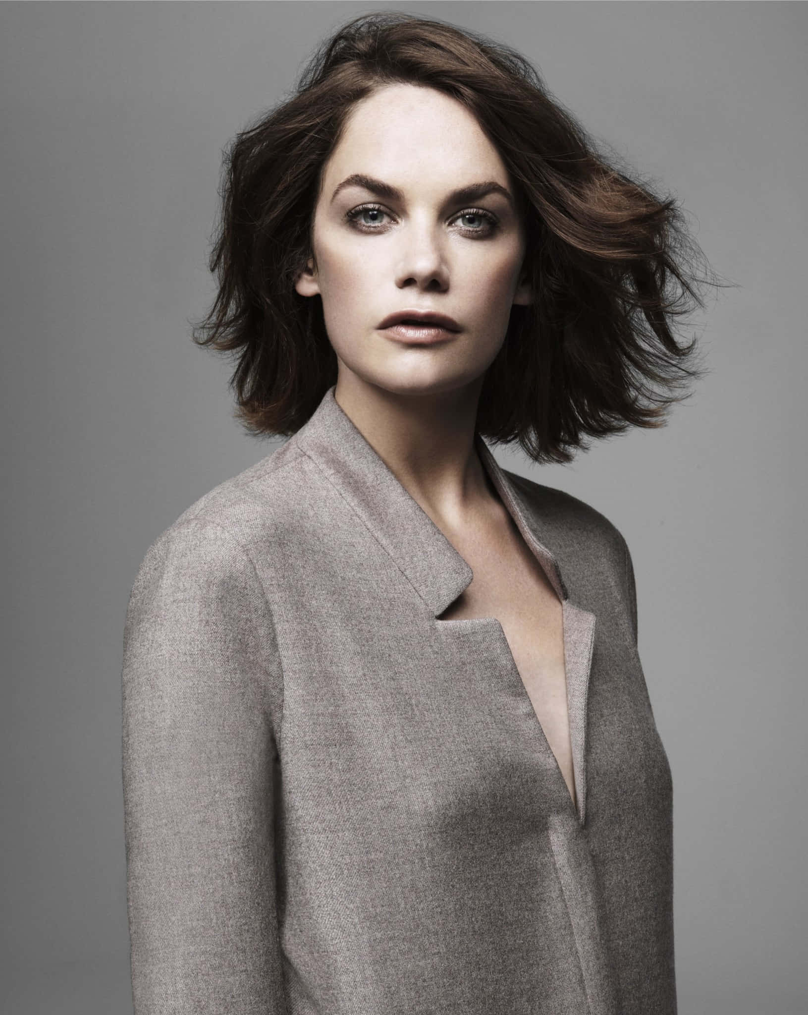 Ruth Wilson 桌布 壁紙 — 一位擁有被風吹亂的棕色頭髮和銳利眼神的女性，在純色攝影棚背景前穿著一件有紋理的灰色西裝外套。