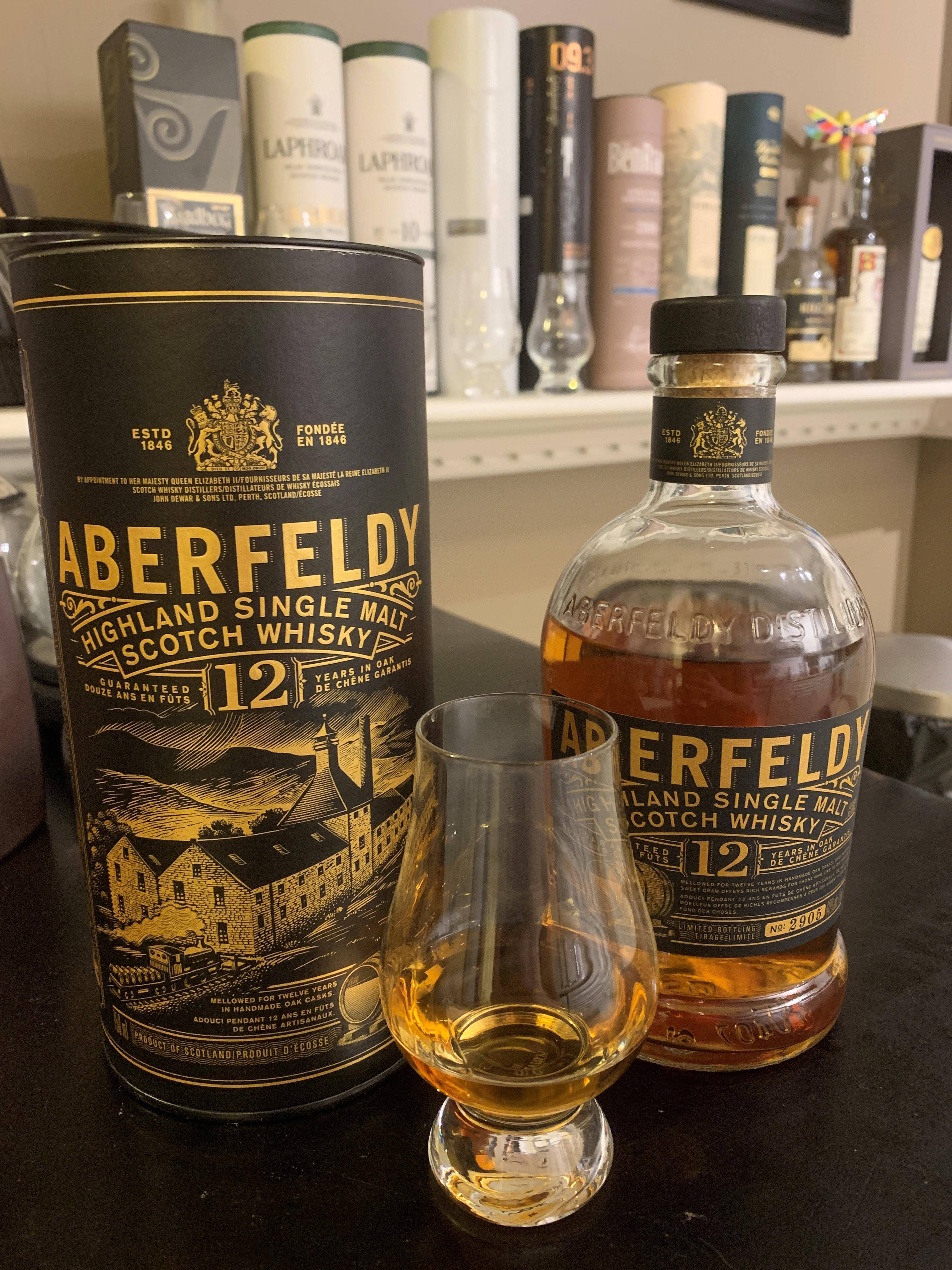 Drink 桌布 壁紙 — Aberfeldy 12年高地單一麥芽蘇格蘭威士忌酒瓶和酒杯放在深色桌子上，背景有品牌包裝和其他烈酒。