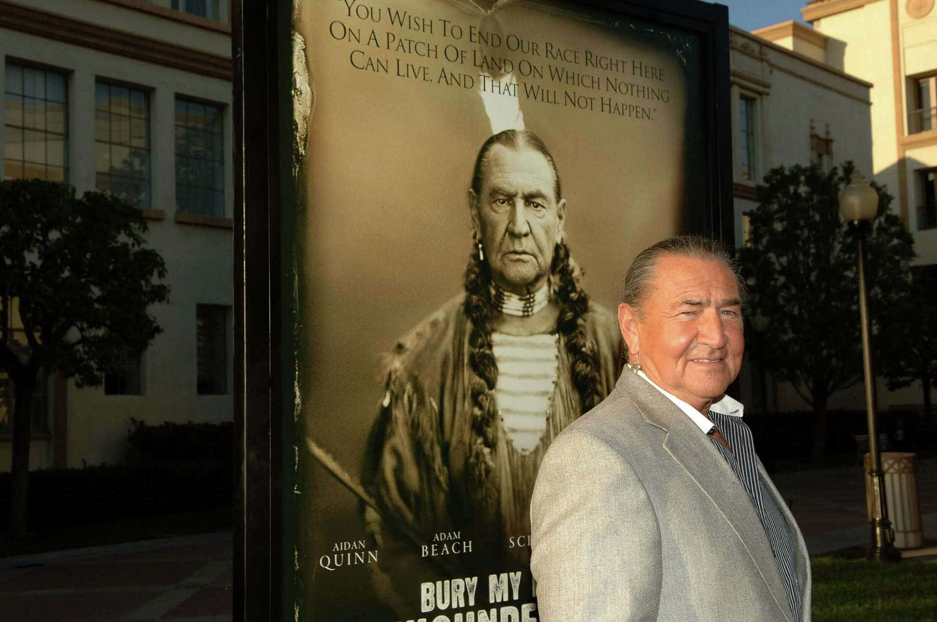  壁紙 — 演員艾丹·奎因站在電影《Bury My Heart at Wounded Knee》的海報旁，海報上是一位編著辮子、穿著傳統服飾的美國原住民男子的肖像。