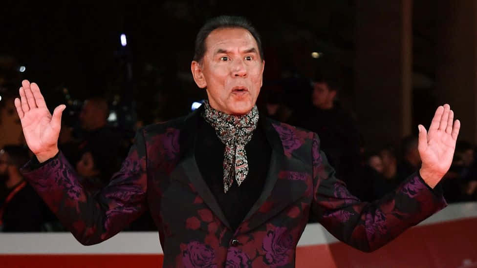  壁紙 — 演員丹尼·特雷喬站在紅地毯上，身穿大膽的紫色花卉西裝，雙手舉起，表現出驚訝或打招呼。