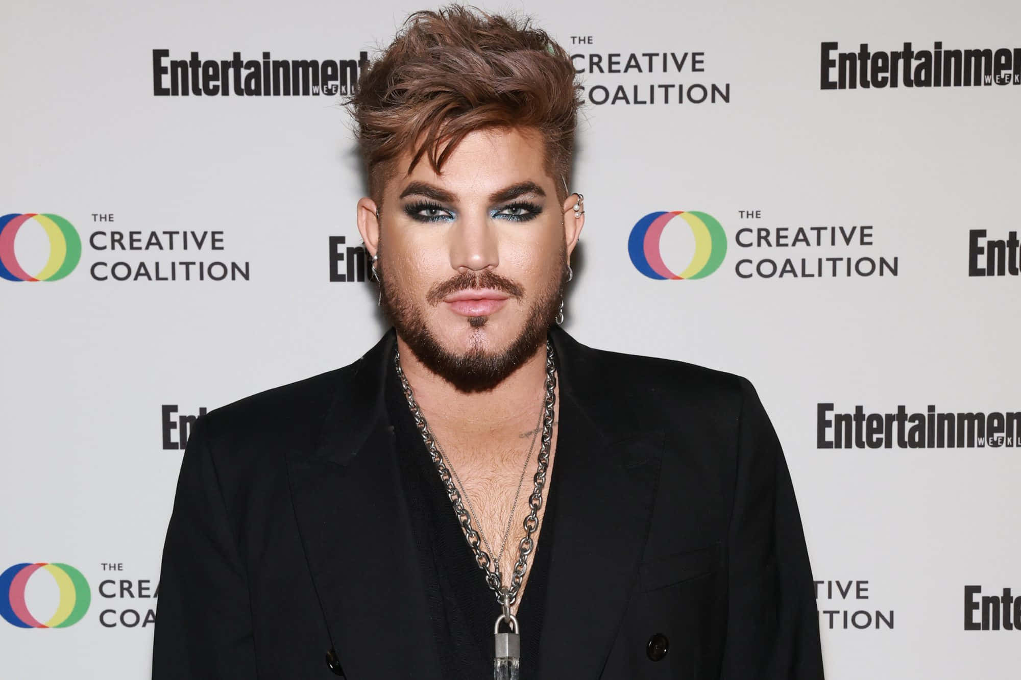 男同志 桌布 壁紙 — Adam Lambert 自信地擺姿勢，身穿黑色西裝，化著大膽的妝容，背景是品牌活動背景。