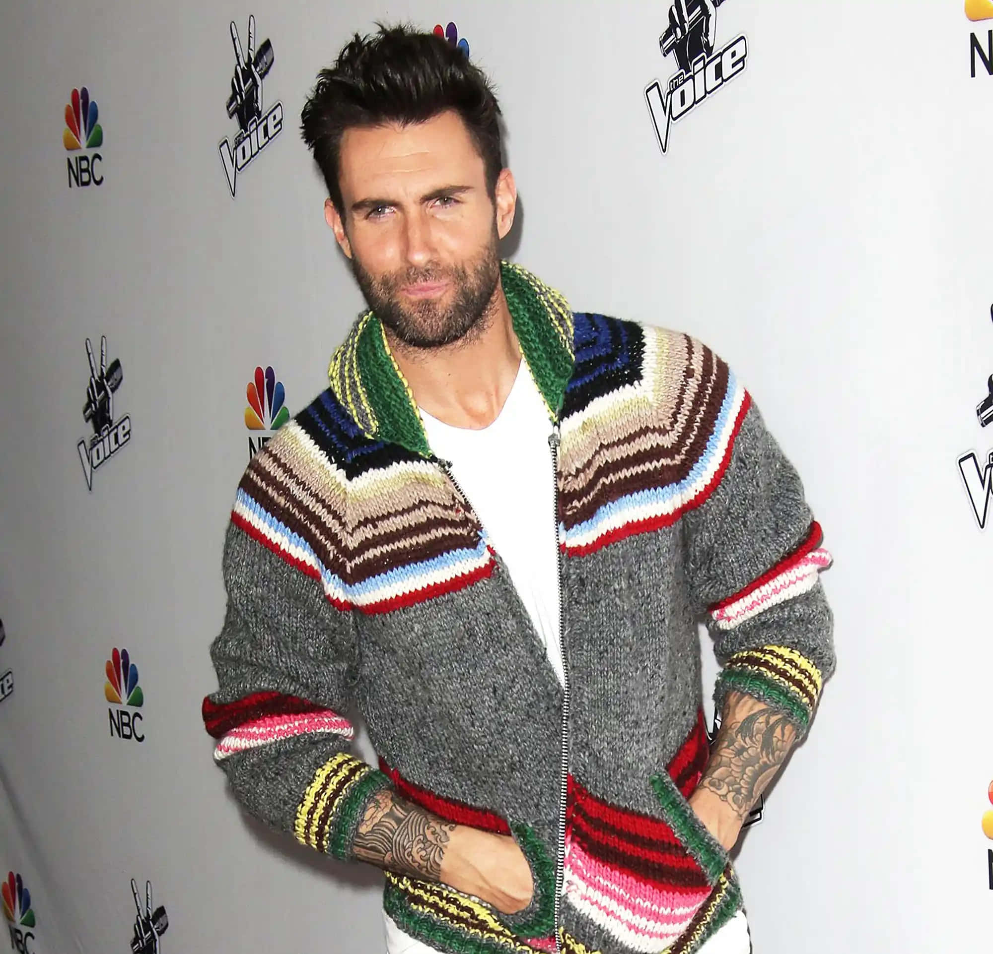 Adam Levine 桌布 壁紙 — Adam Levine 在The Voice活動中自信地擺姿勢，身穿色彩繽紛的條紋針織外套，身後有NBC標誌。