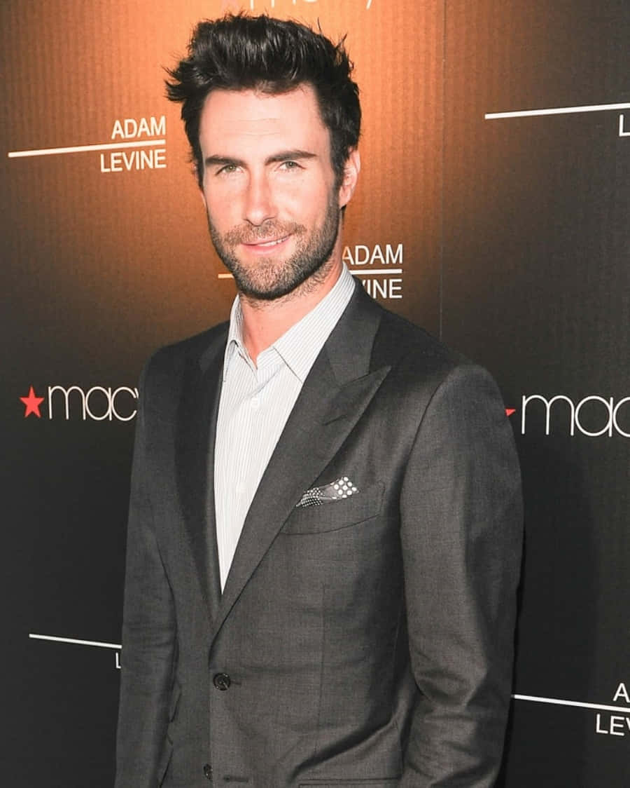 Adam Levine 桌布 壁紙 — Adam Levine 穿著俐落的灰色西裝，在溫暖燈光的品牌活動背景前自信微笑。