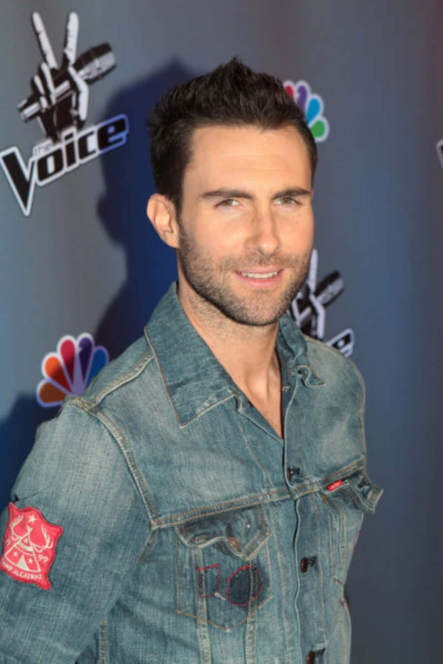 Adam Levine 桌布 壁紙 — Adam Levine 身穿牛仔外套，在背後有NBC標誌的The Voice活動中自信微笑。
