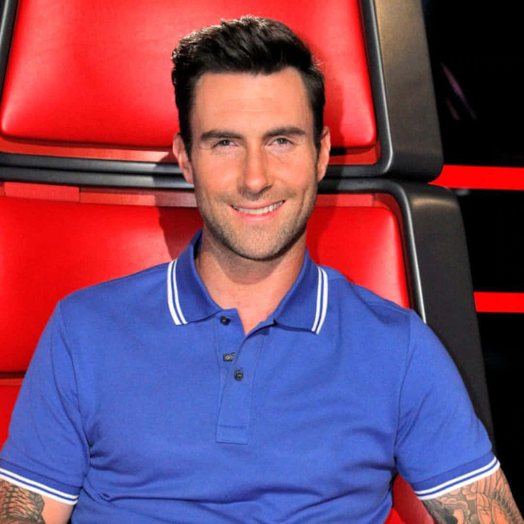 Adam Levine 桌布 壁紙 — Adam Levine 身穿藍色Polo衫，坐在標誌性的紅色評審椅上，溫暖地微笑。