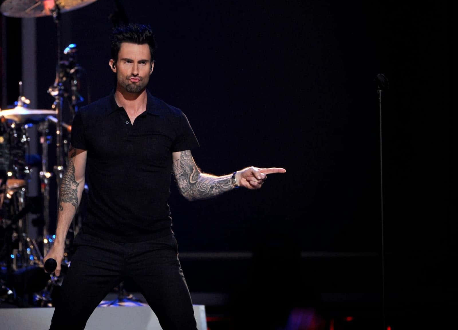 Adam Levine 桌布 壁紙 — Adam Levine 身穿黑色服裝在舞台上表演，向前指著，舞台燈光下可見刺青。