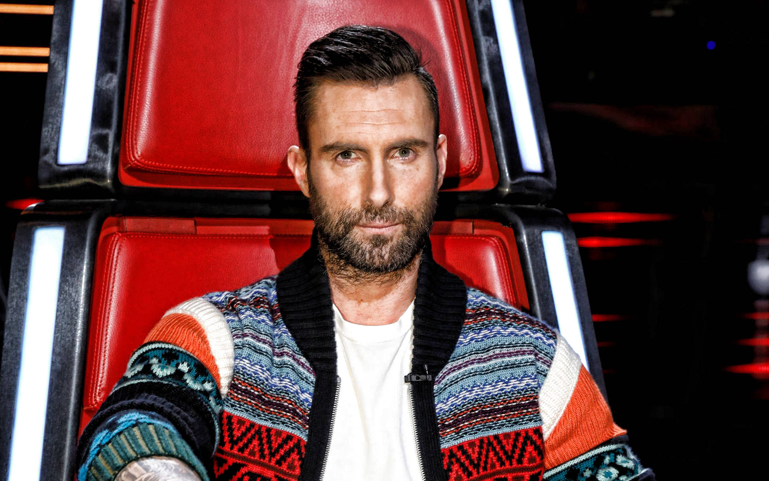 Adam Levine 桌布 壁紙 — Adam Levine自信地坐在《The Voice》標誌性的紅色評審椅上，眼神專注，身穿色彩繽紛的圖案毛衣。