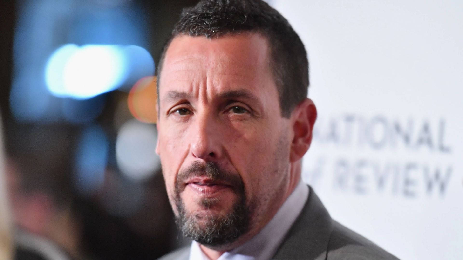 Adam Sandler 桌布 壁紙 — Adam Sandler 在正式活動的特寫肖像，背景有模糊燈光和 'NATIONAL REVIEW'。