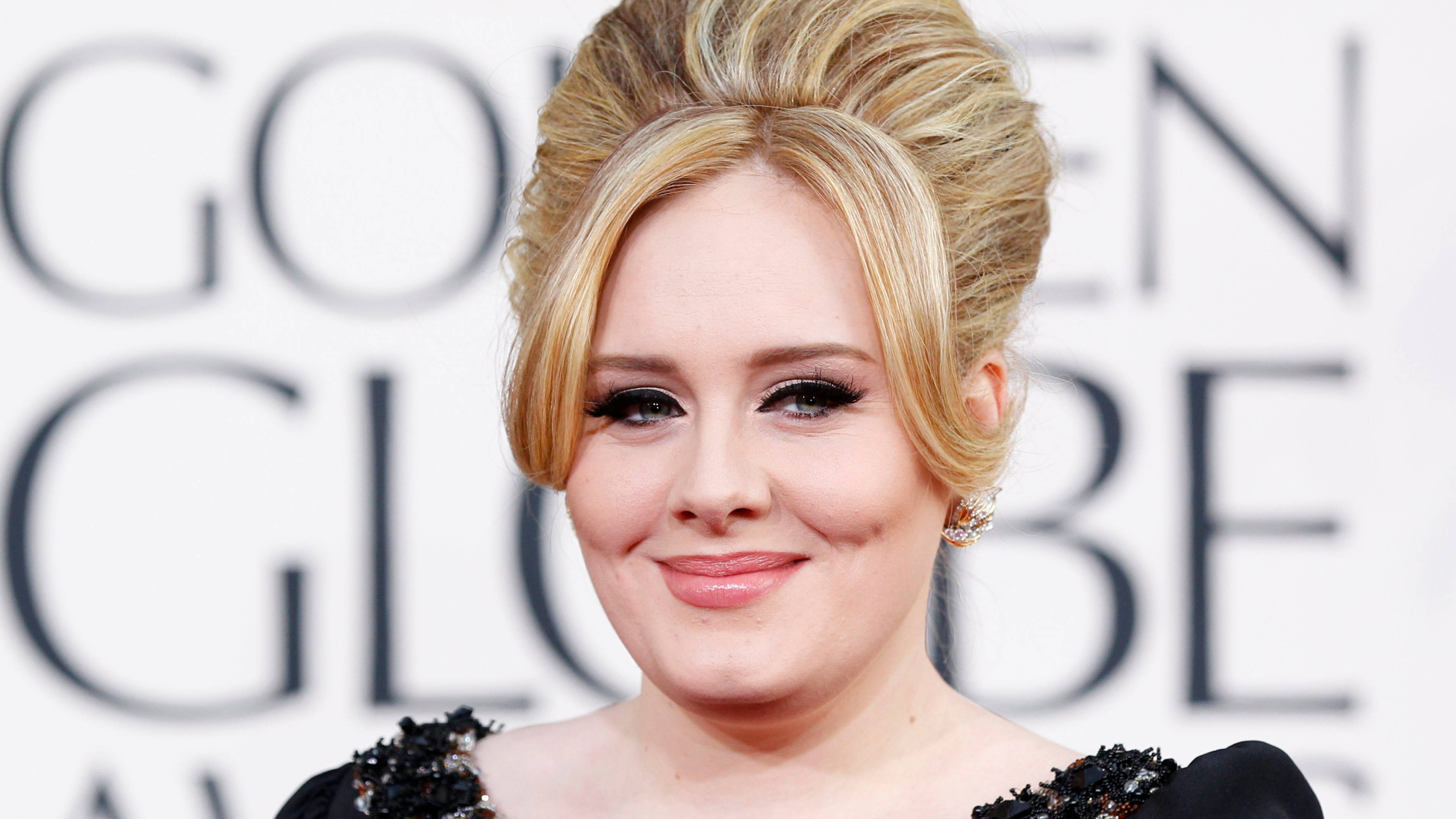 Adele 桌布 壁紙 — Adele 在模糊的金球獎背景下，以優雅的金色盤髮和黑色裝飾連衣裙，溫暖地對著鏡頭微笑。