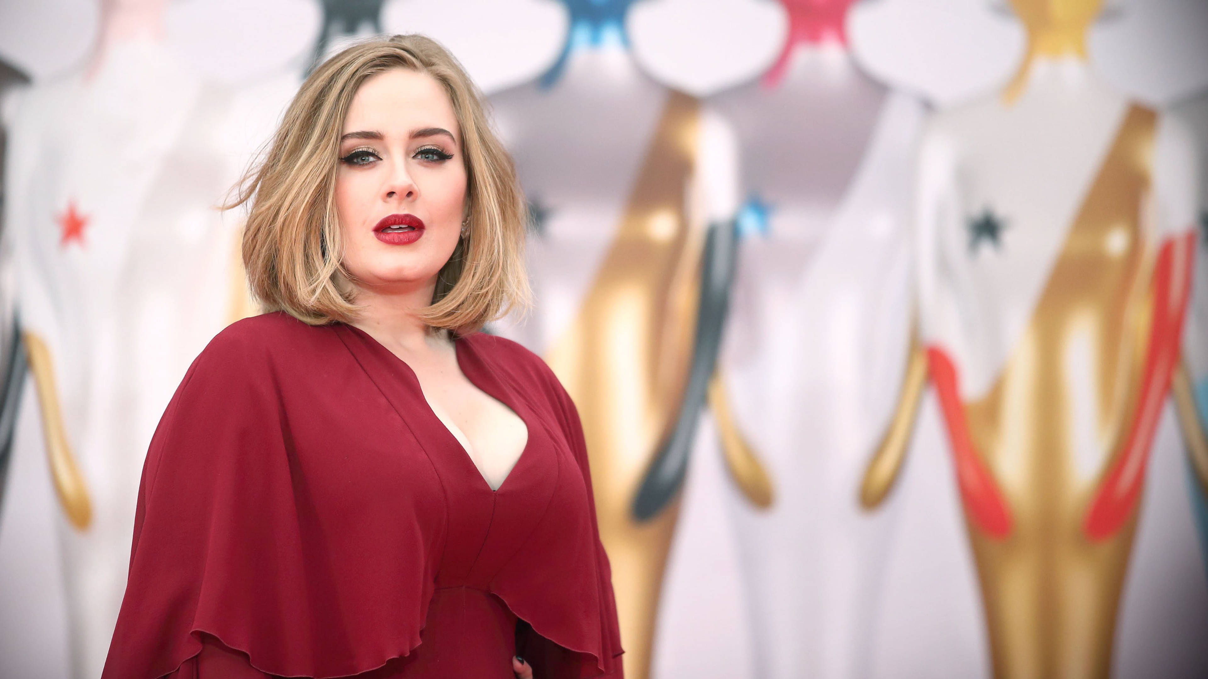 Adele 桌布 壁紙 — Adele 自信地擺姿勢，身穿深紅色長袍，背景是模糊的金色雕像和色彩繽紛的星星。