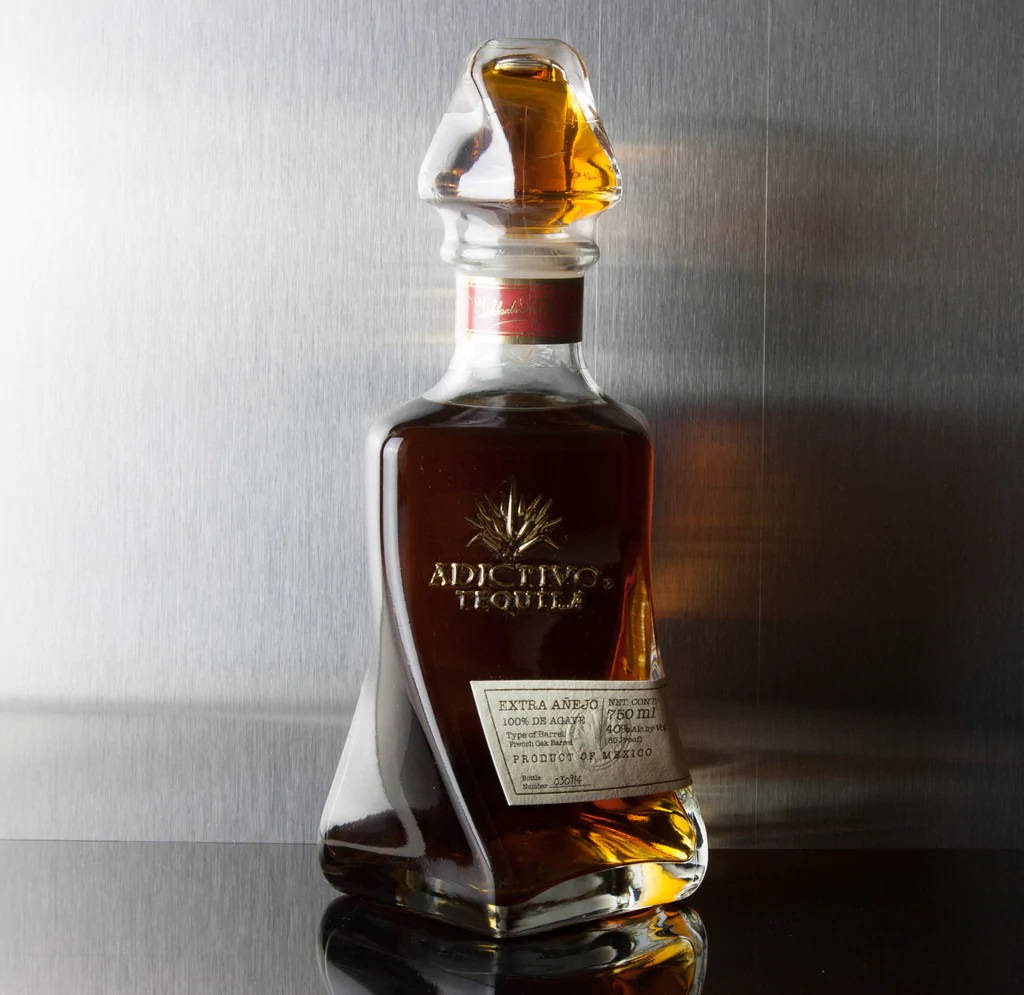 iPhone 桌布 壁紙 — Adictivo Tequila Extra Añejo 的時尚玻璃瓶在拉絲金屬背景下閃耀著琥珀色液體。