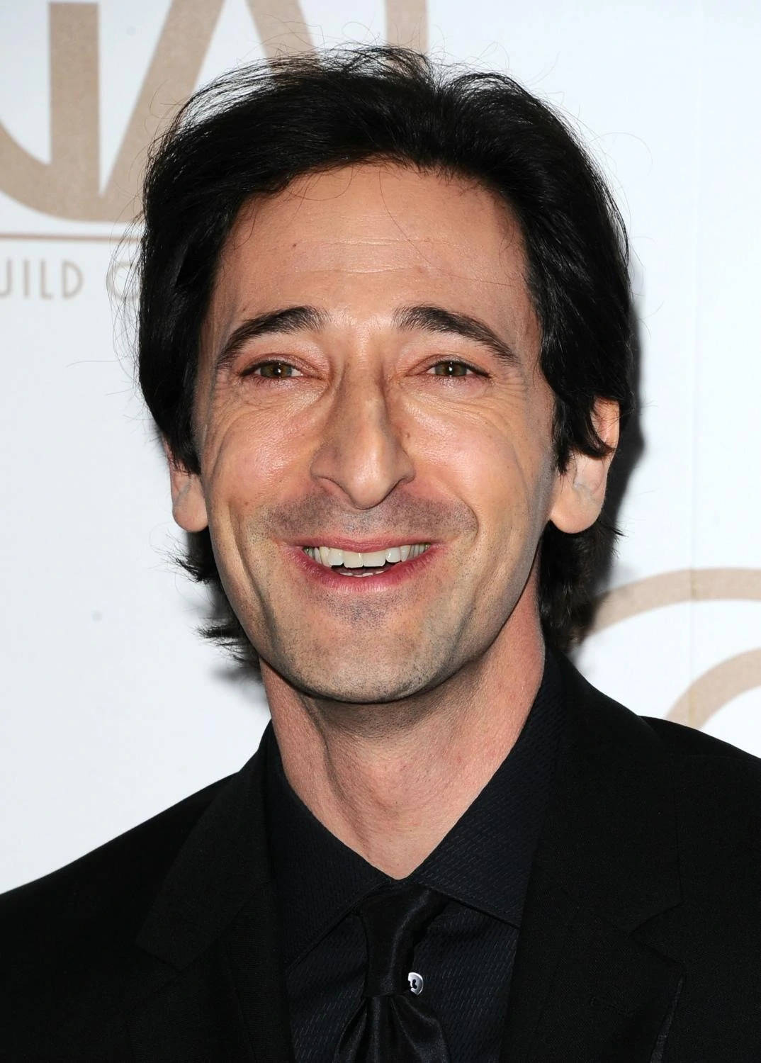 Adrien 桌布 壁紙 — Adrien Brody 身穿黑色西裝，在金色字體的白色活動背景前溫暖地微笑。