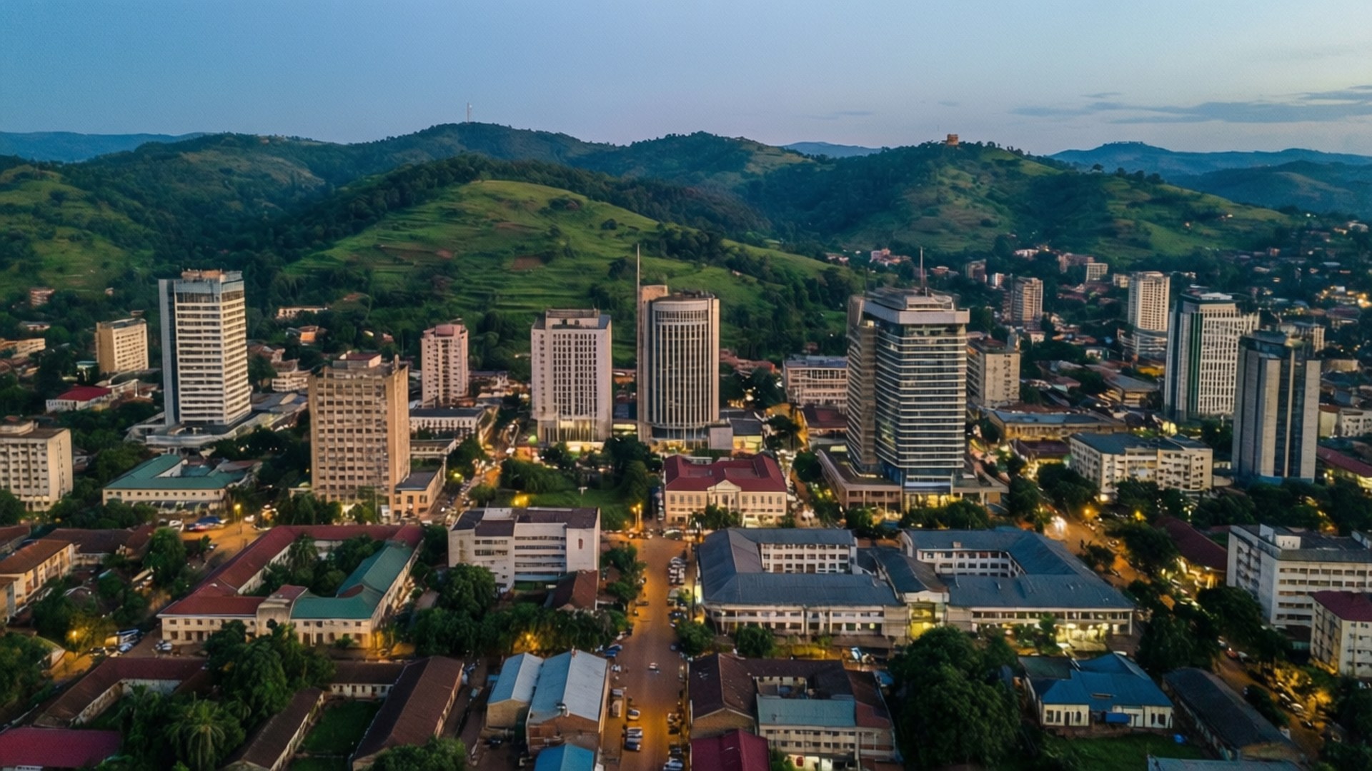 Yaoundé Skyline Evening 壁紙 — 鳥瞰一座坐落在鬱鬱蔥蔥的青山之間的城市，暮色中街道燈火通明，現代建築林立。