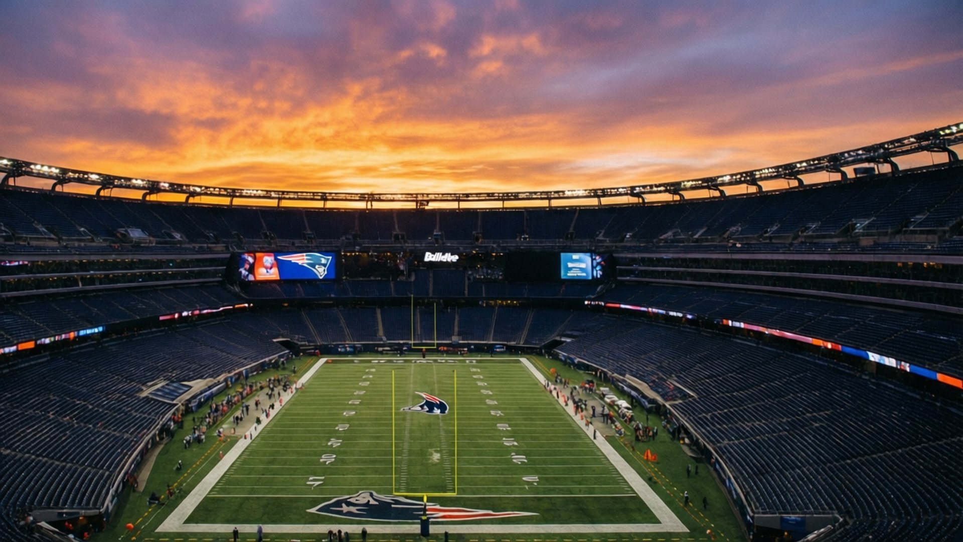 Gillette Stadium Sunset 壁紙 — 日落時足球場的鳥瞰圖，空藍色座位上方的鮮豔橙色雲朵和帶有球隊標誌的綠色場地。