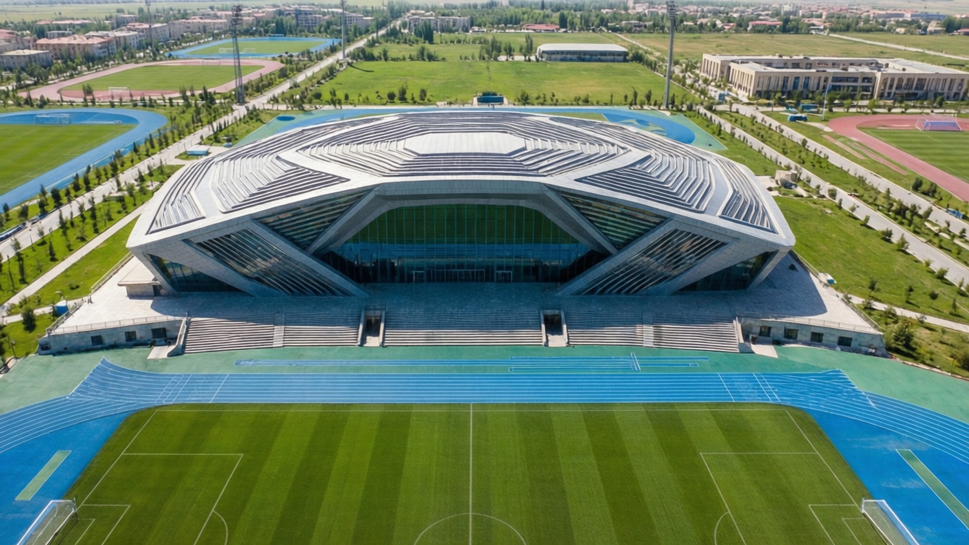 Sports Complex Aerial 壁紙 — 未來主義體育館的鳥瞰圖，幾何屋頂，周圍環繞著綠色田野和藍色跑道。