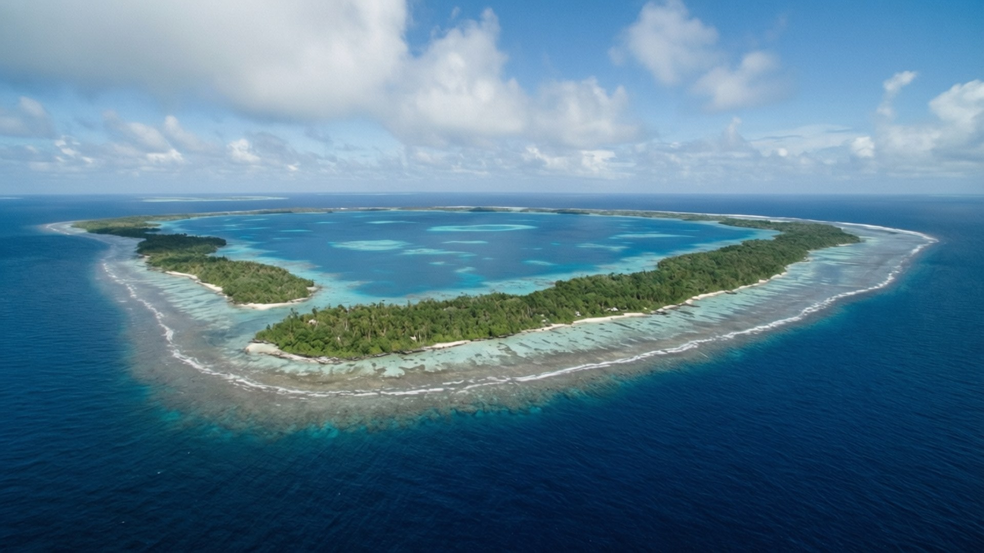 Remote Atoll Wilderness 壁紙 — 在部分多雲的天空下，一個鬱鬱蔥蔥的綠色環礁被綠松石潟湖和深藍色海洋環繞的鳥瞰圖。
