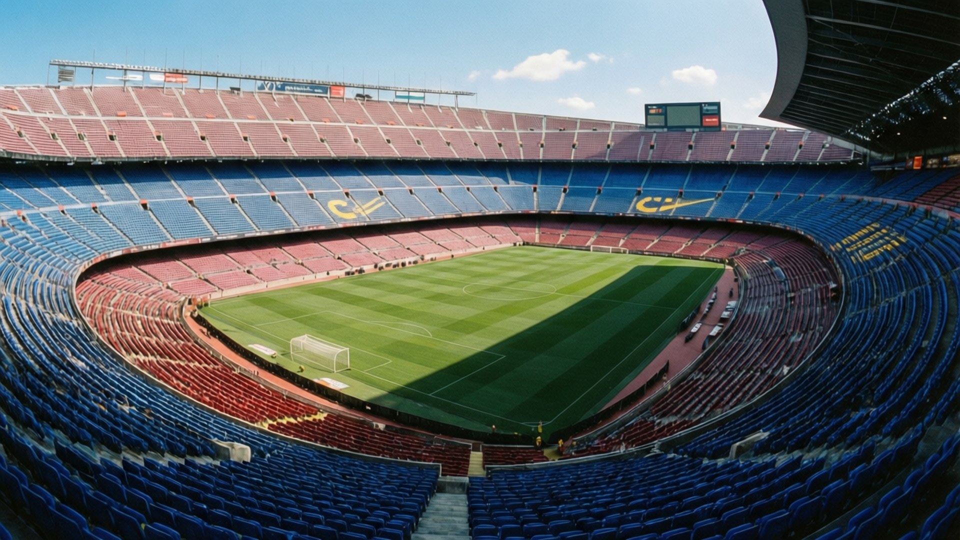 Camp Nou Aerial View 壁紙 — 晴朗天空下，一個巨大空蕩的足球場鳥瞰圖，充滿活力的綠色球場被藍色和紅色座位環繞。