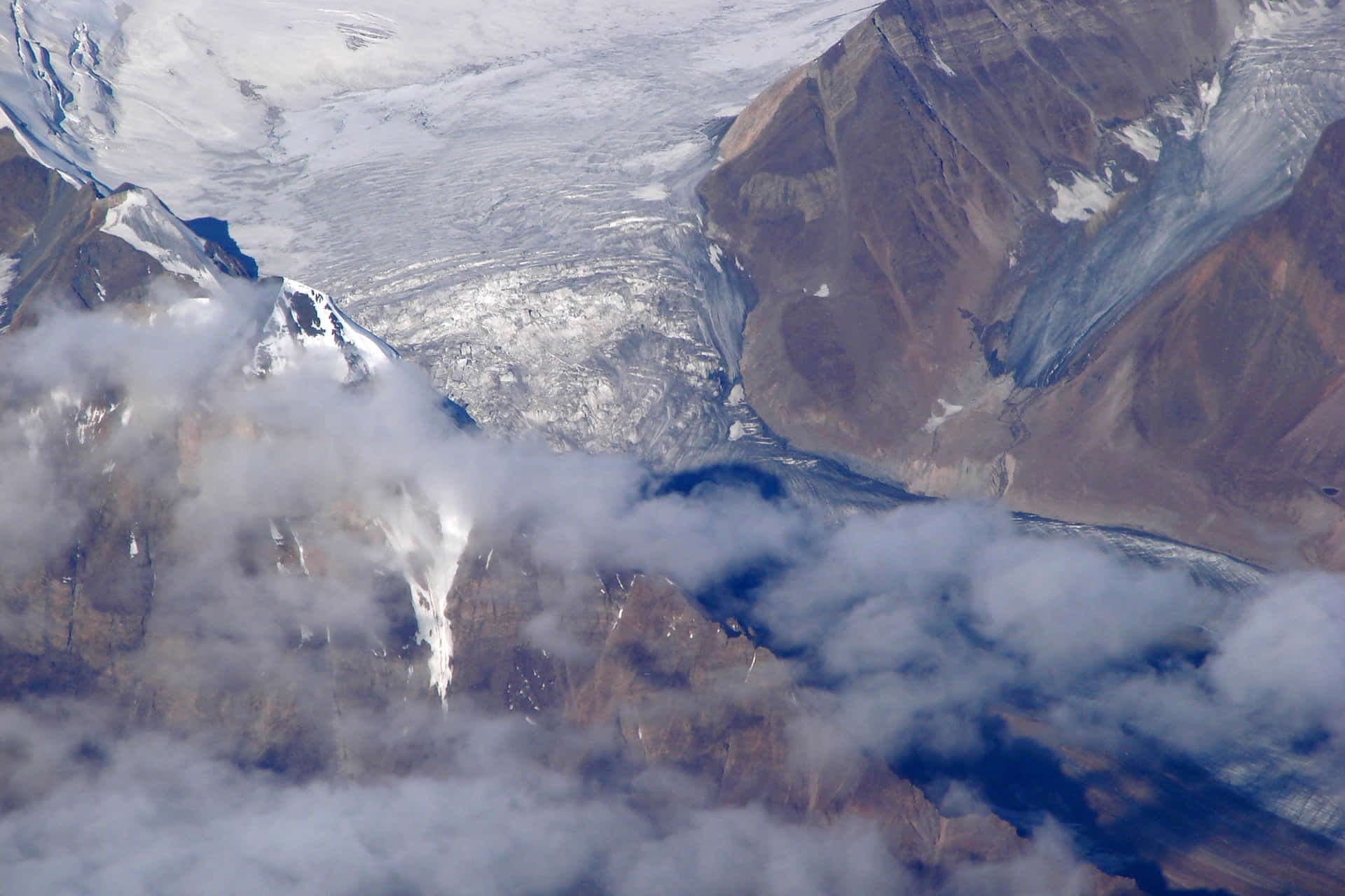  壁紙 — 從高空俯瞰，巨大冰川切割崎嶇山脈地形，部分被飄浮雲層遮蔽。