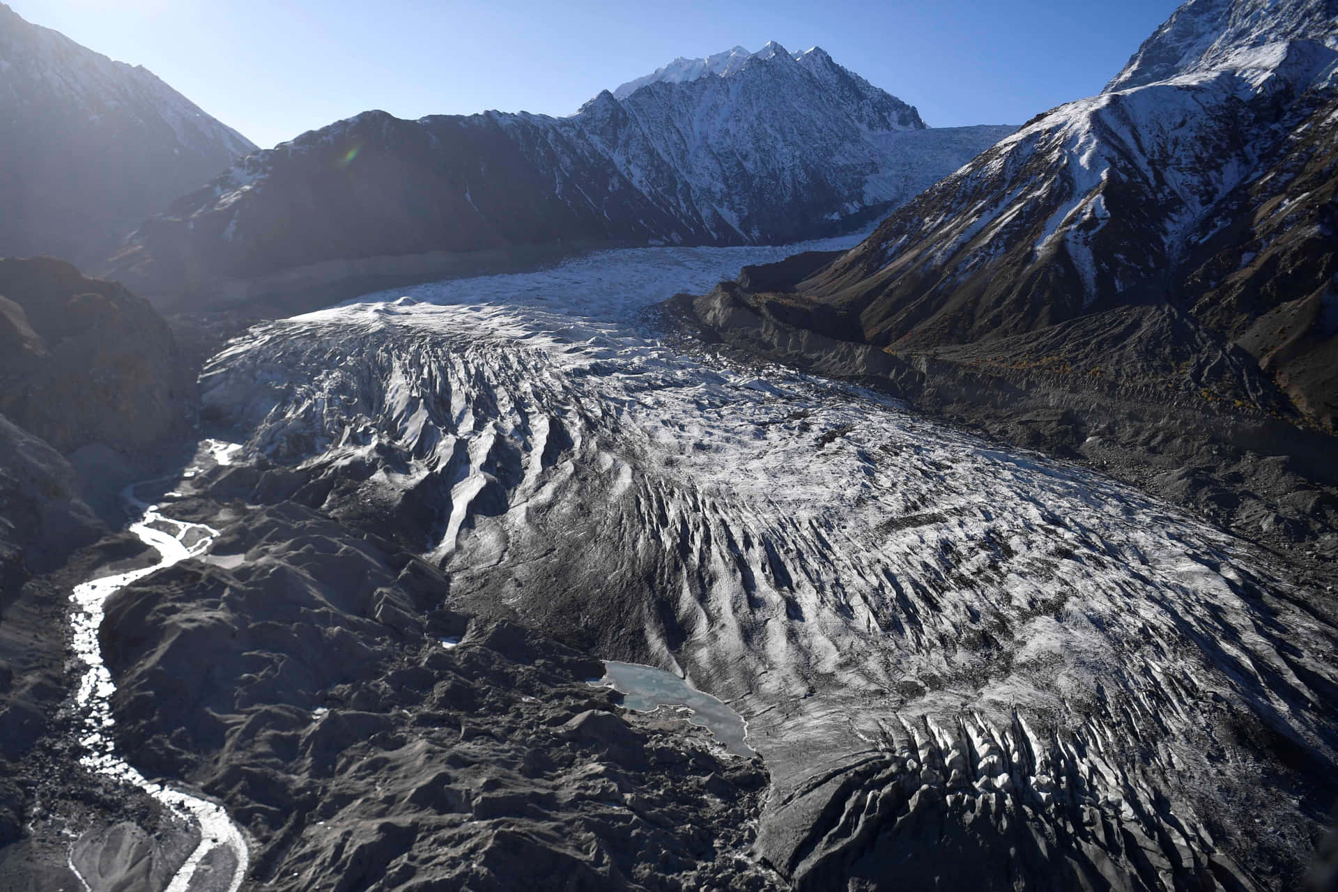  壁紙 — 從高空俯瞰，一座巨大的冰川在崎嶇的雪山間蜿蜒流淌，天空湛藍清澈。