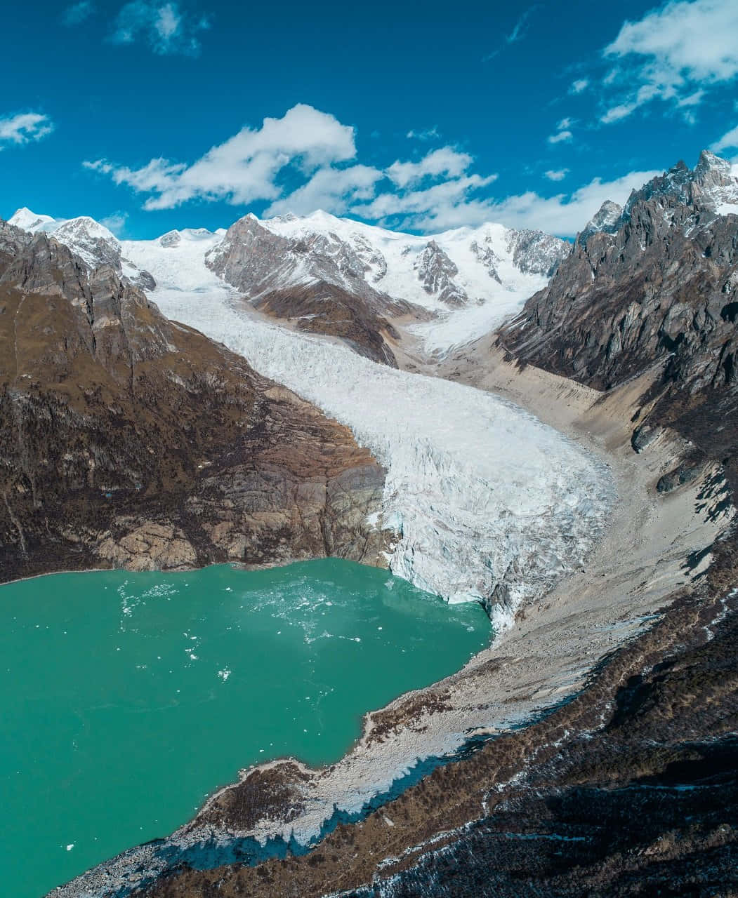  壁紙 — 從高空俯瞰，巨大的冰川從白雪皚皚的山峰流入充滿活力的綠松石冰川湖，上方是明亮的藍天，點綴著幾朵雲彩。