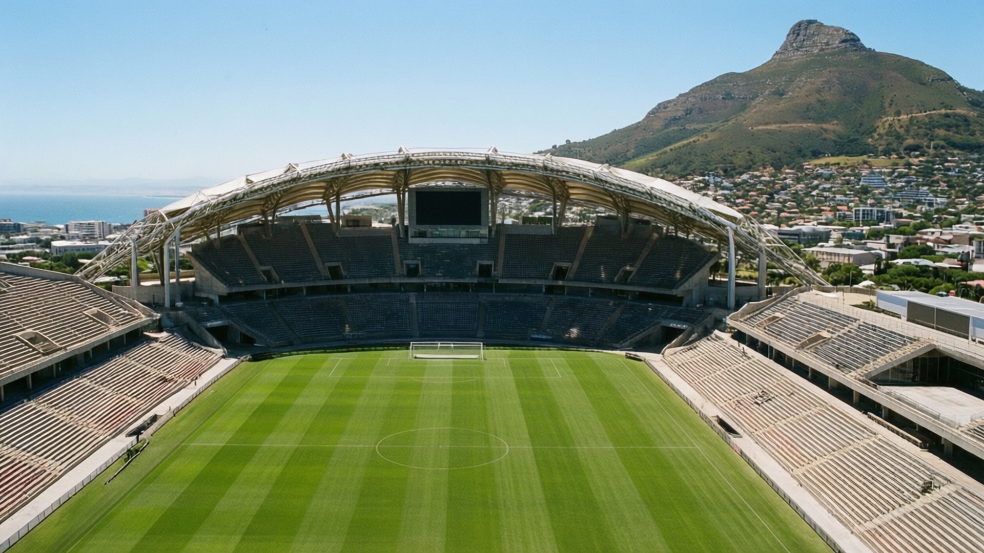 Cape Town Stadium Aerial 壁紙 — 現代足球場的鳥瞰圖，擁有鮮綠色草地和空蕩蕩的看台，背景是晴朗的藍天和一座山。