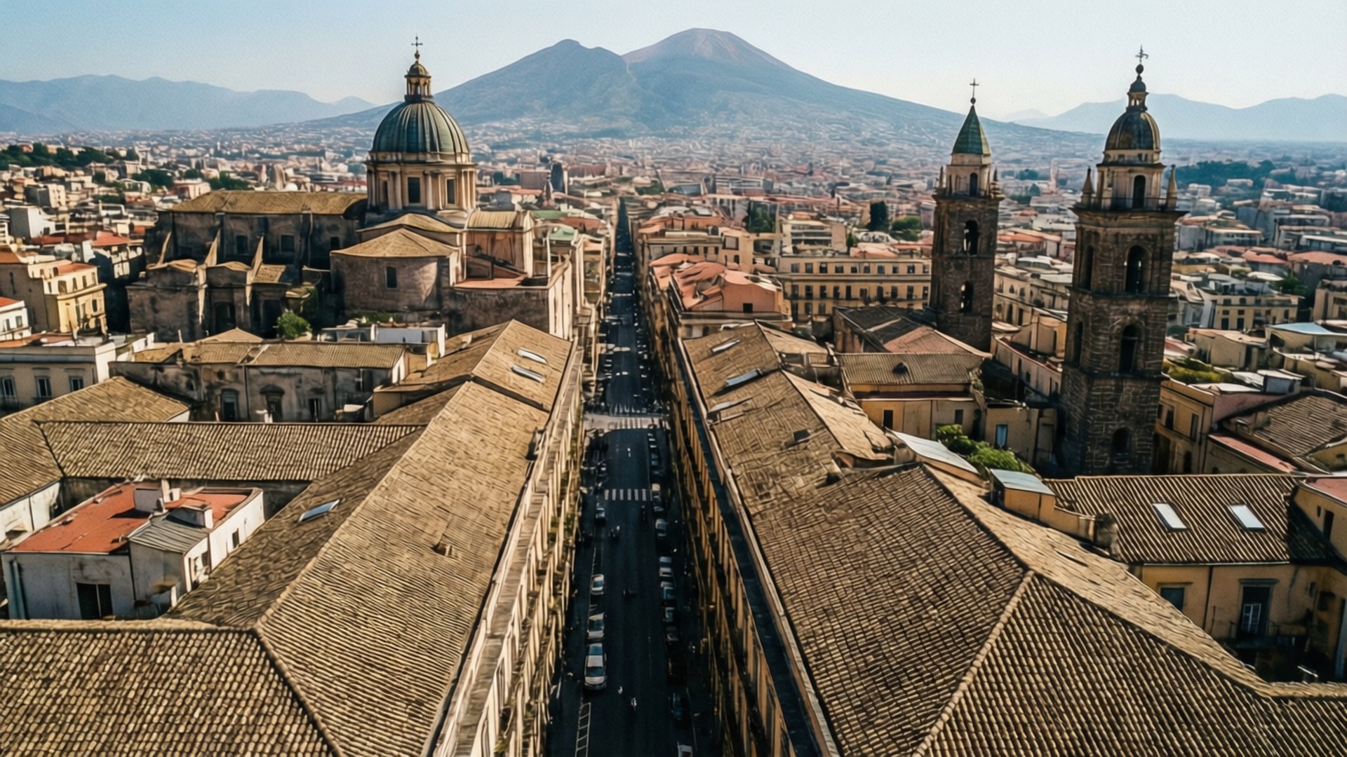 Naples Rooftop Aerial 壁紙 — 陽光普照的歐洲城市街道鳥瞰圖，兩旁是陶土屋頂的建築物，遠處有一座火山聳立。