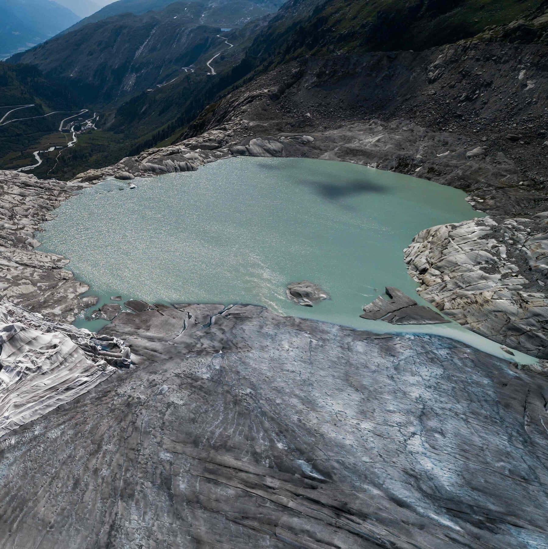  壁紙 — 崎嶇山地中碧綠冰川湖的航拍視角，遠處可見蜿蜒的道路。