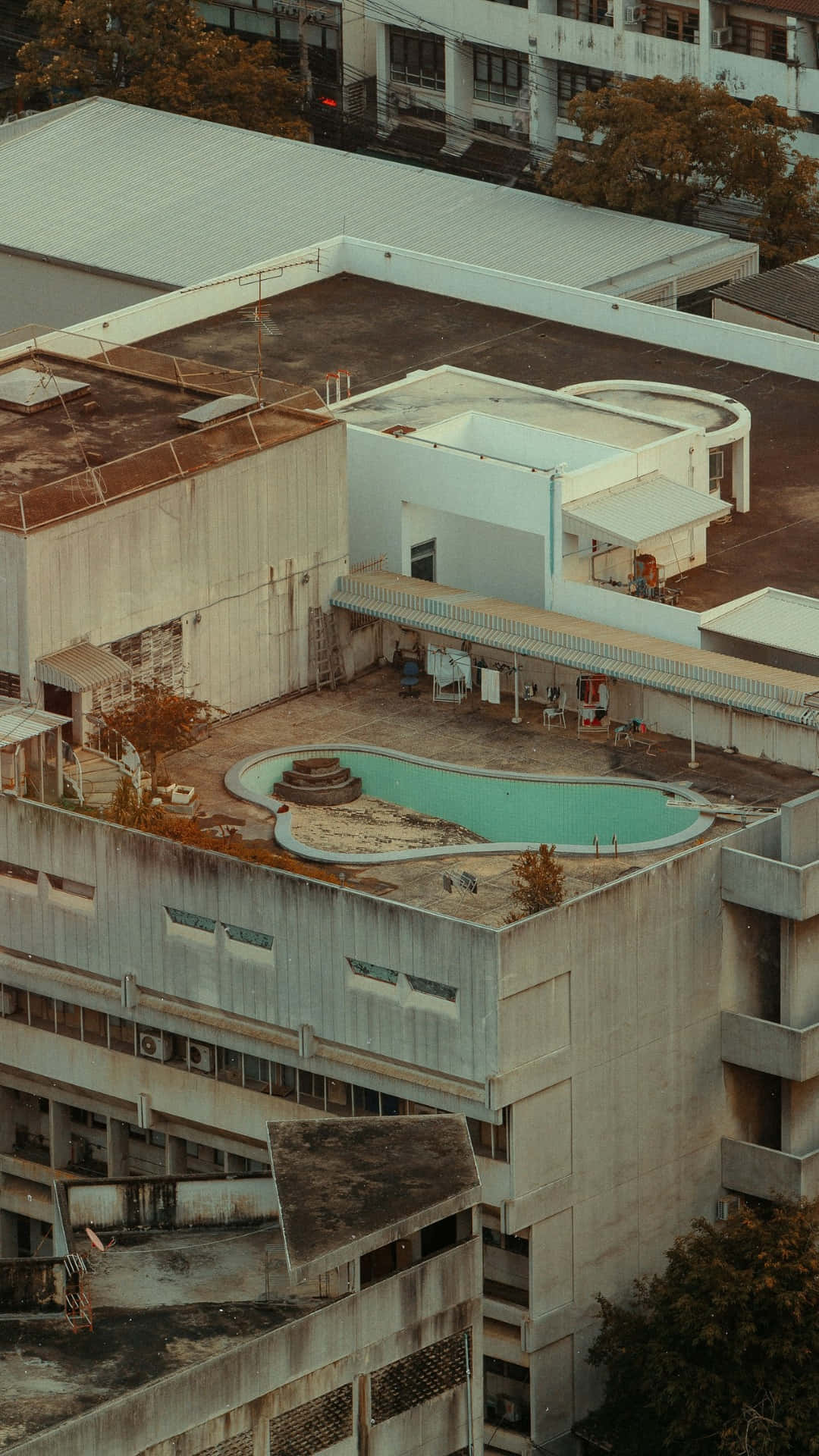  壁紙 — 從高空俯瞰，一個風化的屋頂泳池被混凝土建築和秋天的樹木環繞，沐浴在溫暖的金色光線中。