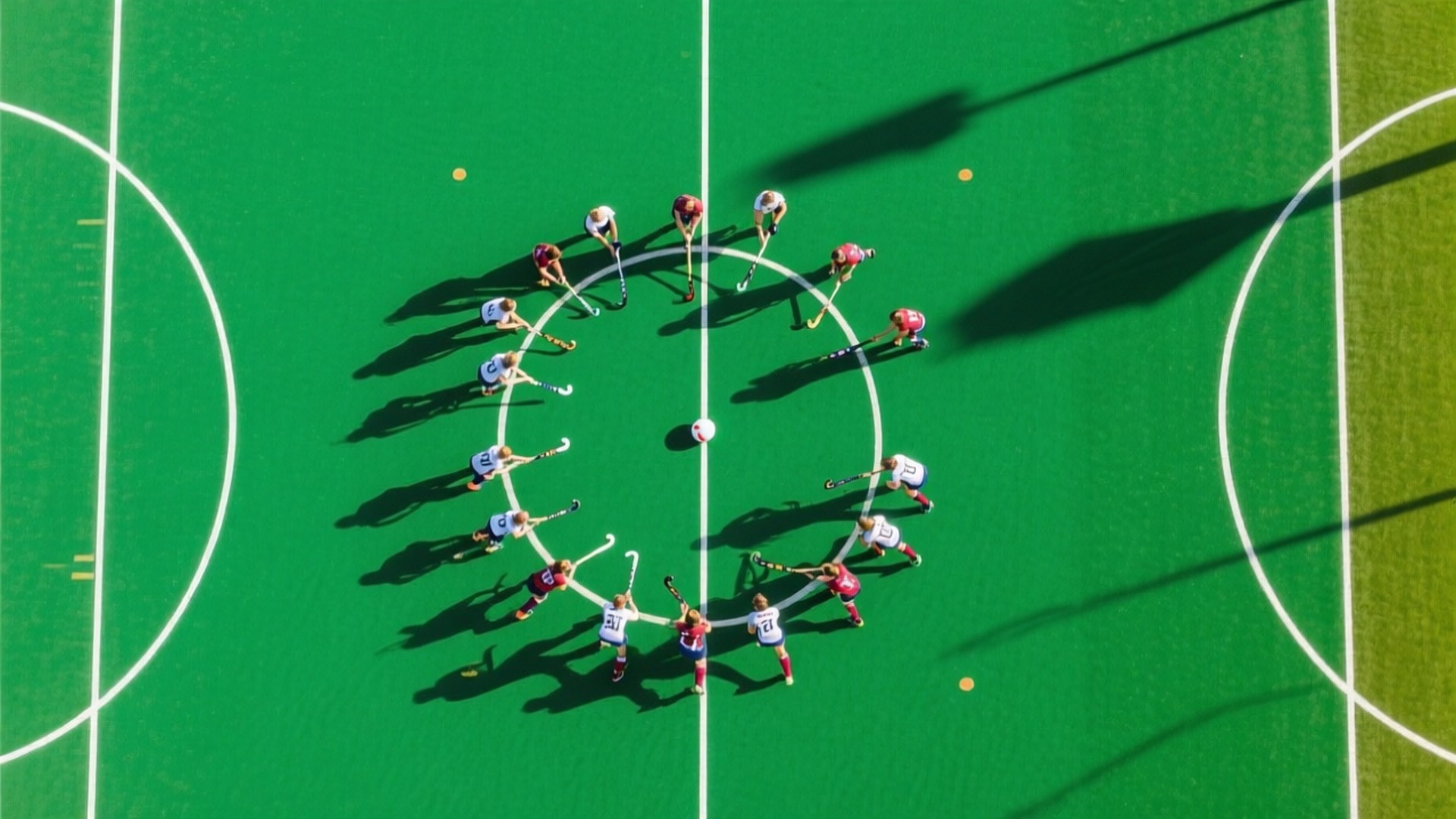 Match from Above 壁紙 — 從高空俯瞰，身穿紅白球衣的曲棍球球員在翠綠的球場上圍成一個圓圈，留下長長的影子。