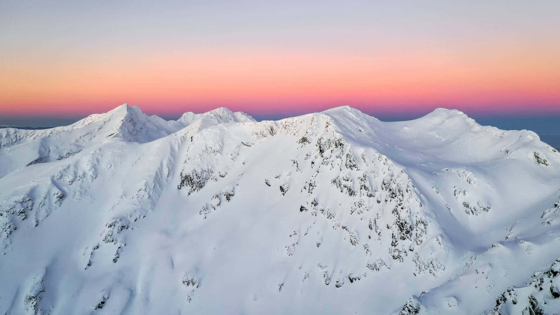  壁紙 — 白雪覆蓋的山峰在晴朗天空下沐浴在柔和粉紅和橙色日出色調中的航拍視圖。