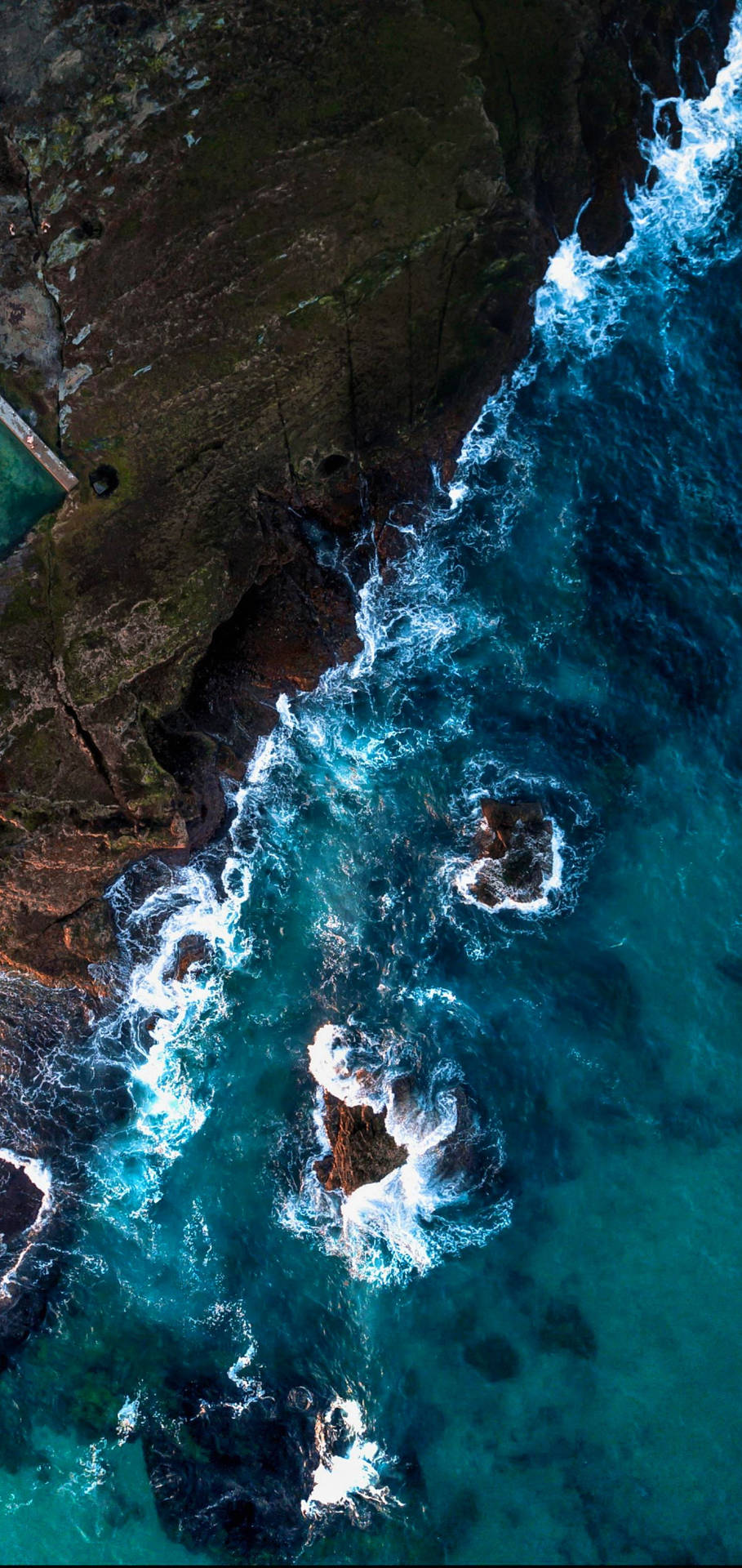藍綠色的海浪從上方拍打黑色的岩石懸崖。 — iPhone X 壁紙