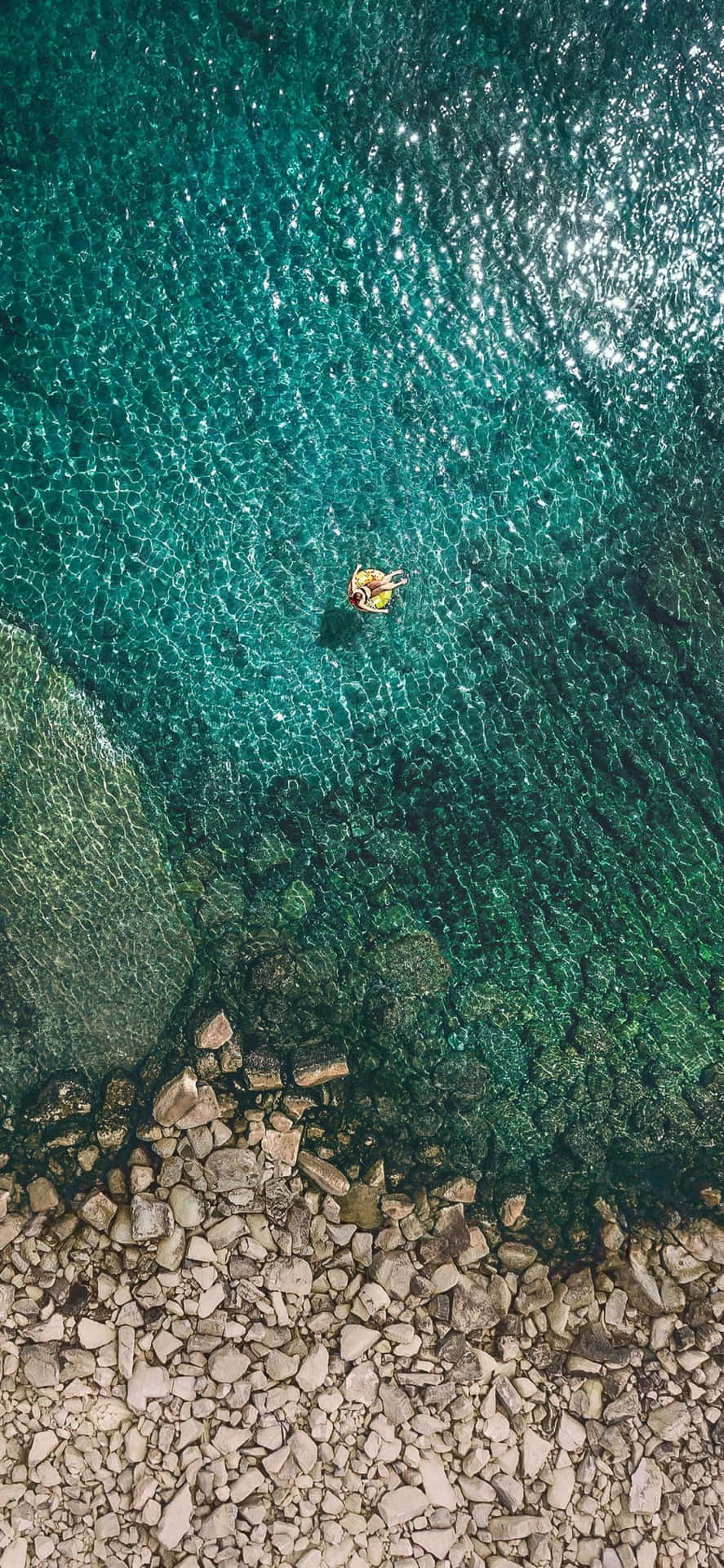 人漂浮在黃色充氣圈上，身處清澈碧綠的海水中，靠近岩石海岸。 — iPhone 11 Pro Max 壁紙
