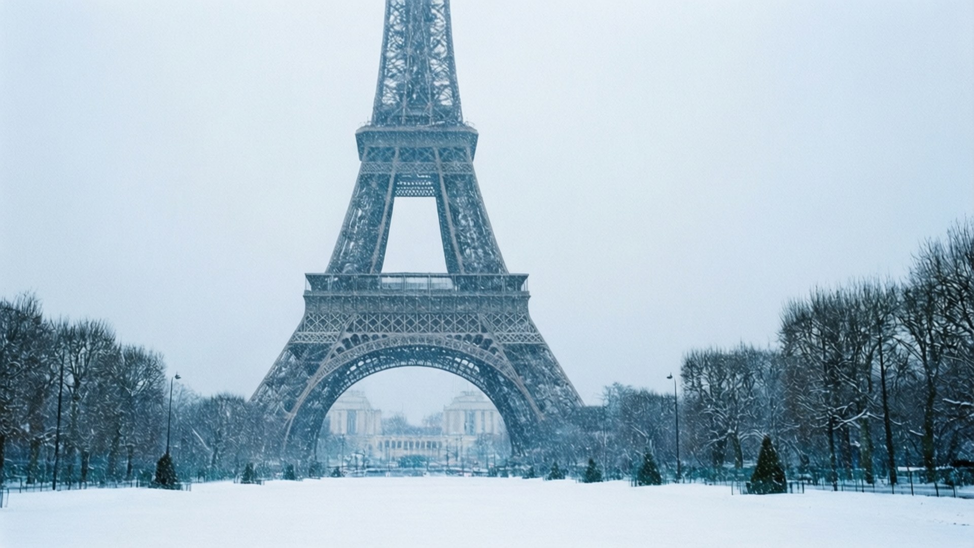 Winter Wonderland Eiffel 壁紙 — 艾菲爾鐵塔在白雪覆蓋的寧靜冬季景觀中高聳，光禿禿的樹木排列在冰凍的地面上。
