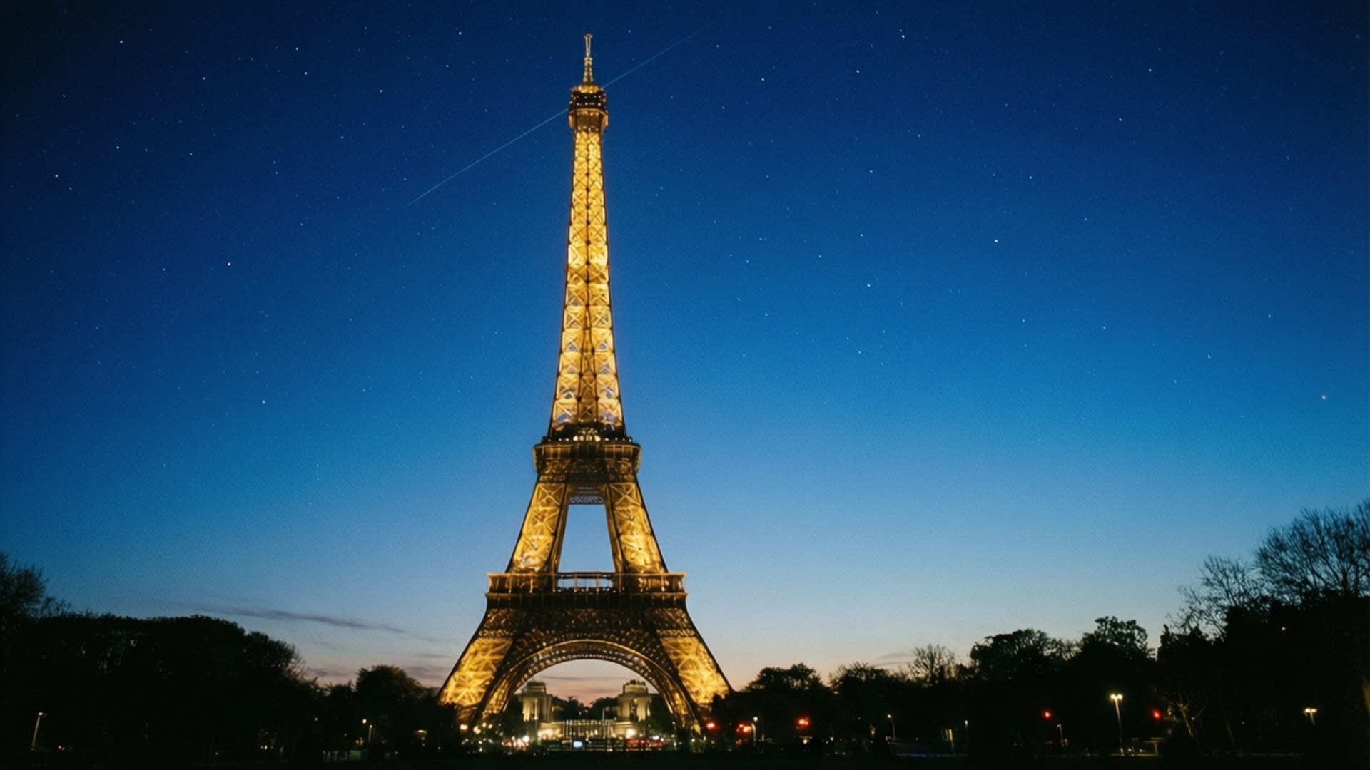 Blue Hour Eiffel 壁紙 — 艾菲爾鐵塔在深藍色暮色天空中溫暖地發光，天空點綴著星星和微弱的飛機尾跡。